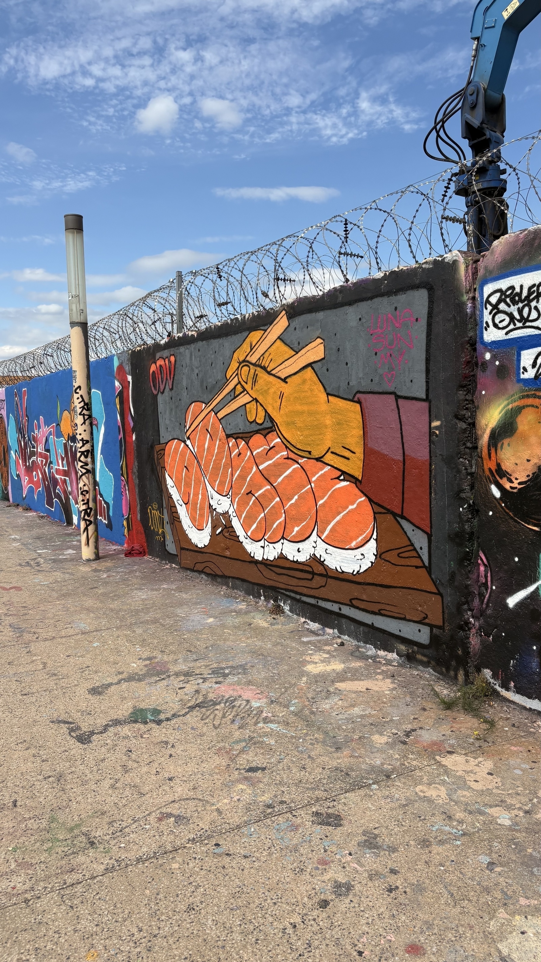 A vibrant mural depicts a hand using chopsticks to pick up pieces of sushi, set against a graffiti-covered wall with barbed wire above.