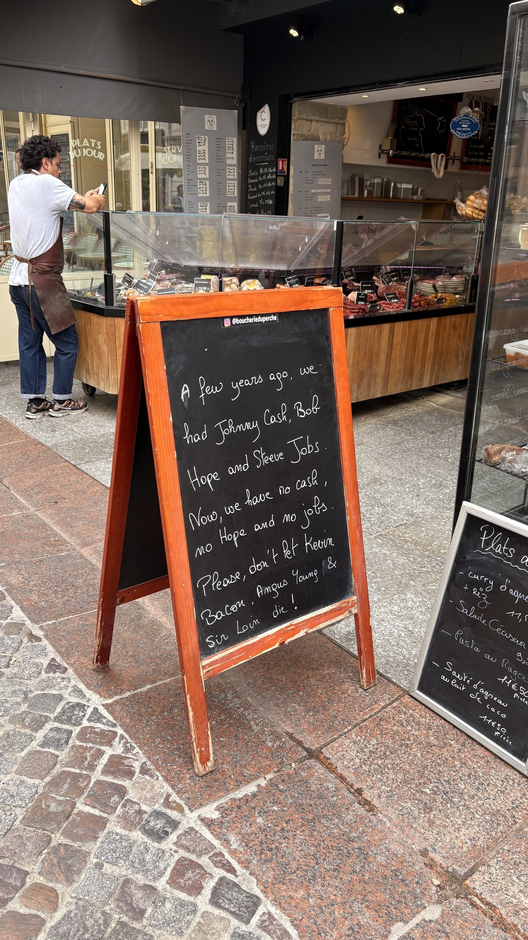 A chalkboard outside a deli humorously references loss of hope, cash, jobs, and bacon, while a person works inside.