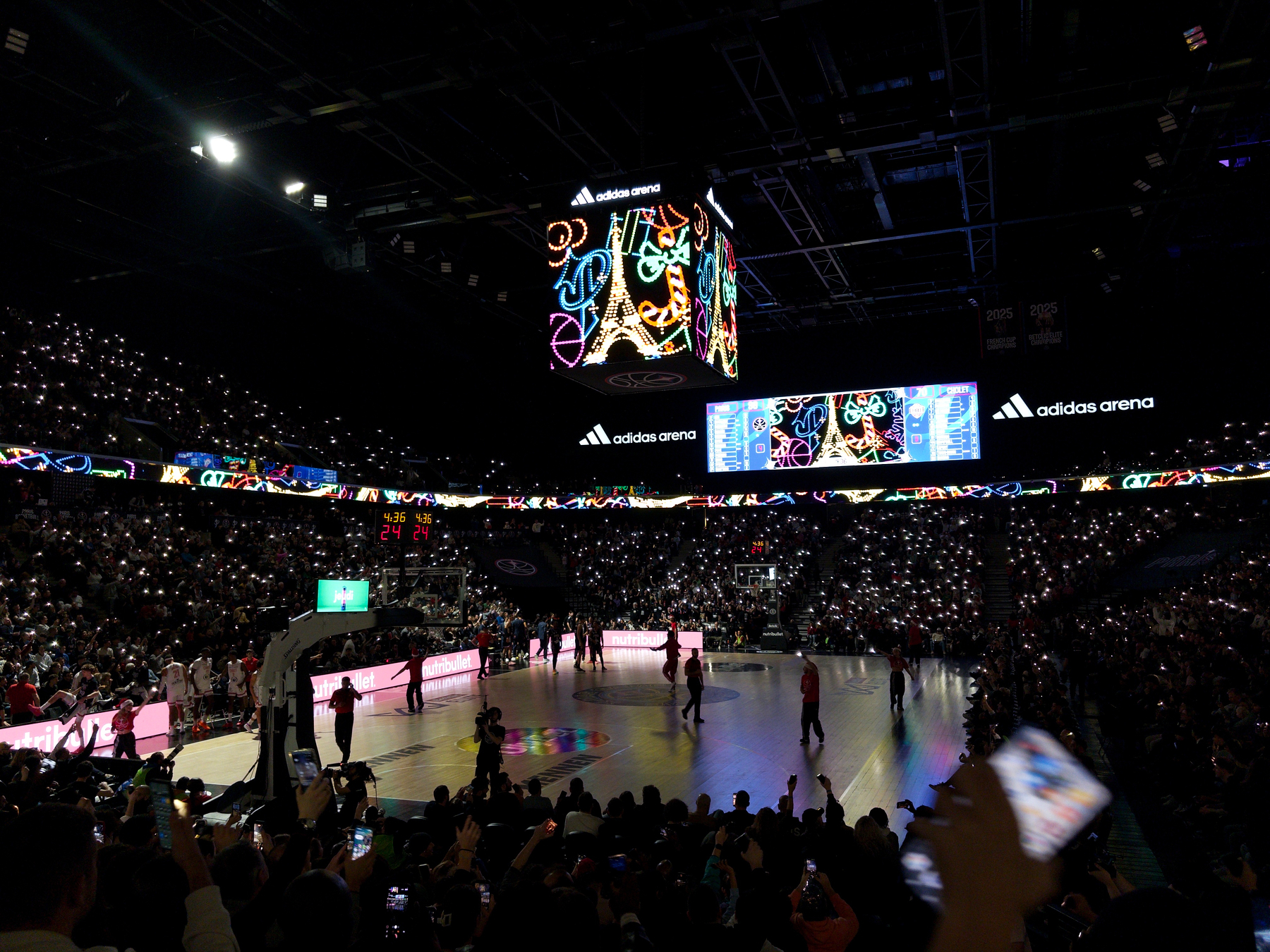 A lively basketball game is taking place in a packed arena with colorful lights and an illuminated scoreboard.