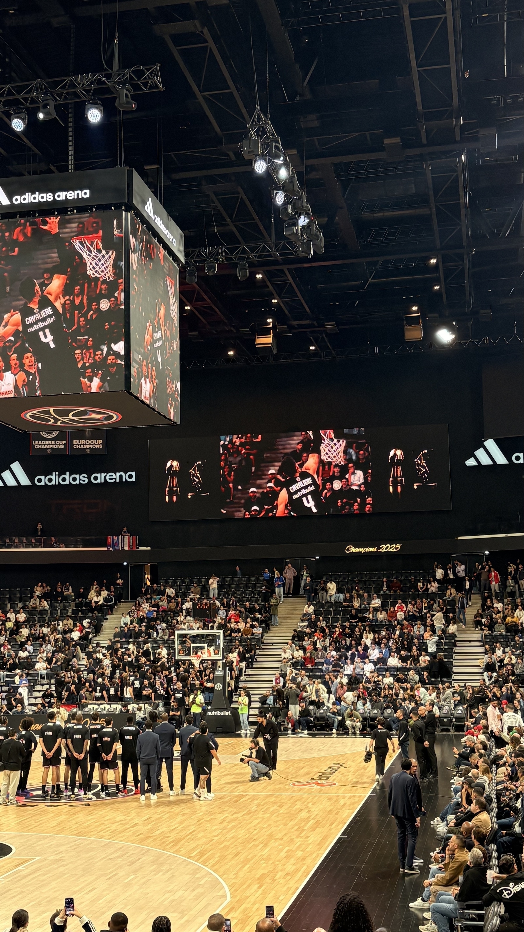 A basketball game is taking place in an arena with spectators watching and multiple large screens displaying the action.