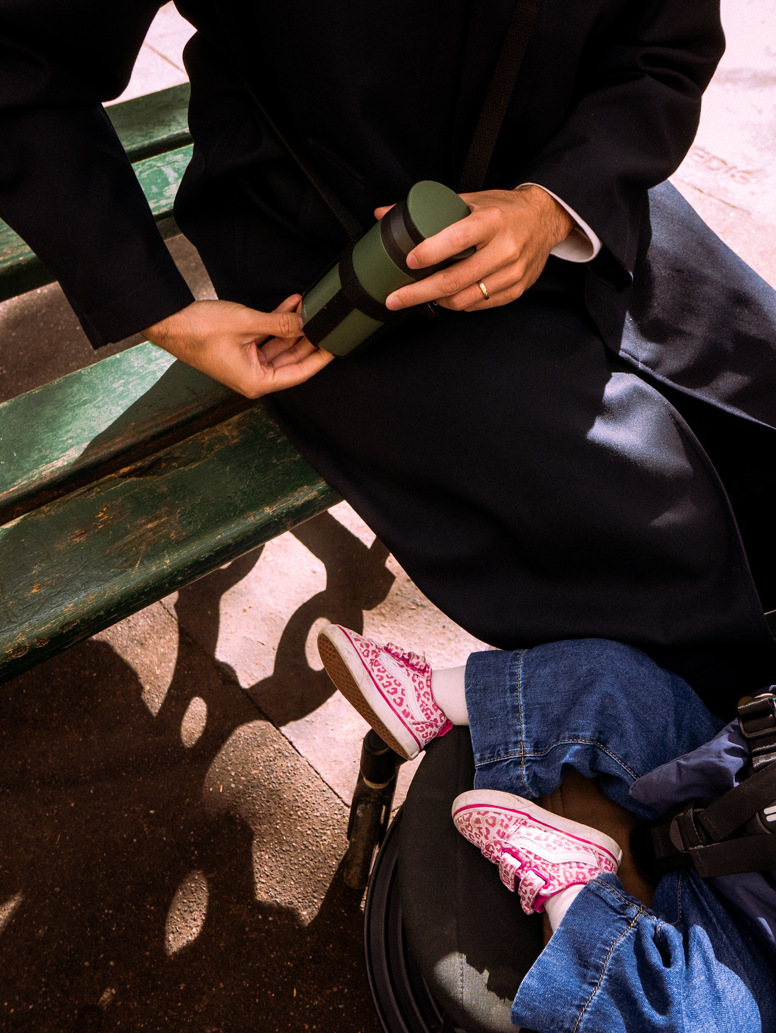 A person is sitting on a bench, holding a thermos, with a child wearing pink shoes in a stroller nearby.