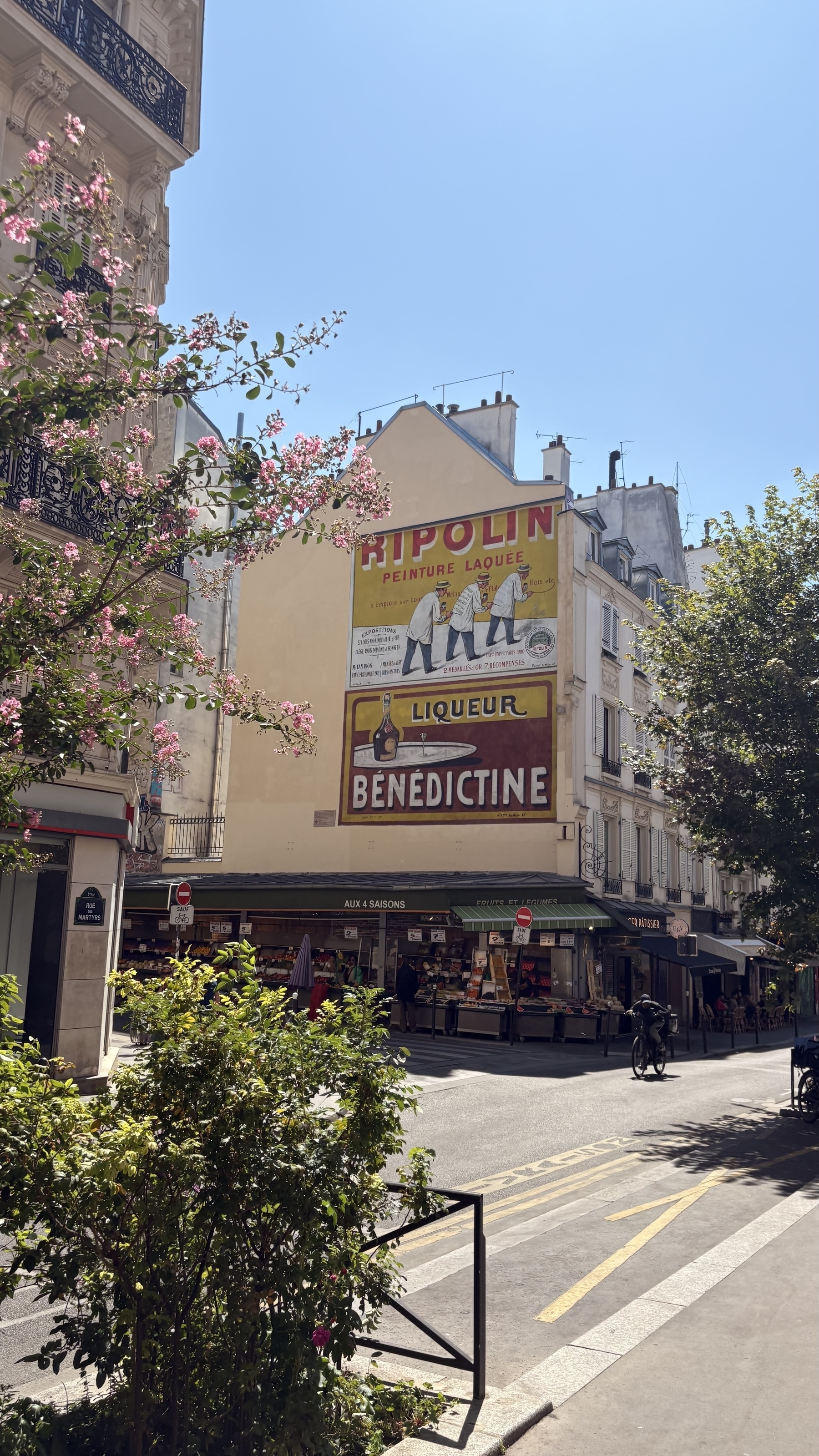 A street scene in a sunny city features a vintage advertisement on a building facade, lush greenery, and a small market or kiosk.