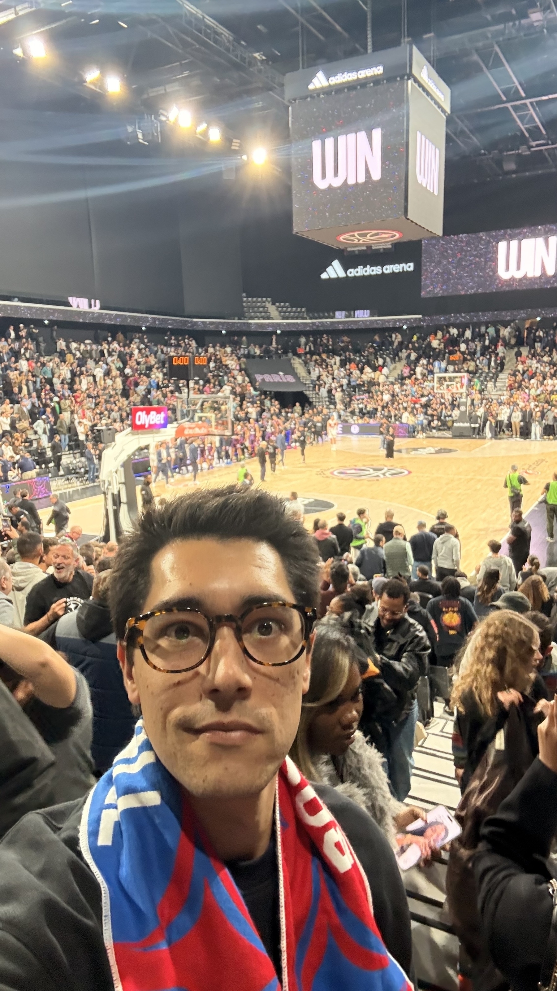 A person wearing glasses and a scarf takes a selfie in a crowded basketball arena with a game underway and WIN displayed on the screen above the court.