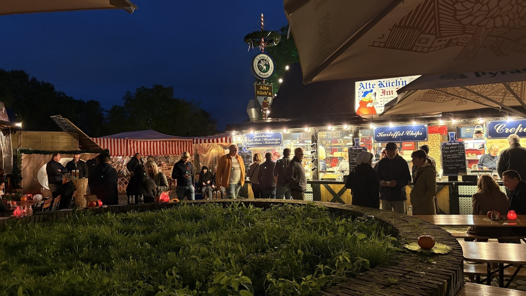People are gathered at an outdoor market or festival in the evening, with food stalls and illuminated canopies.