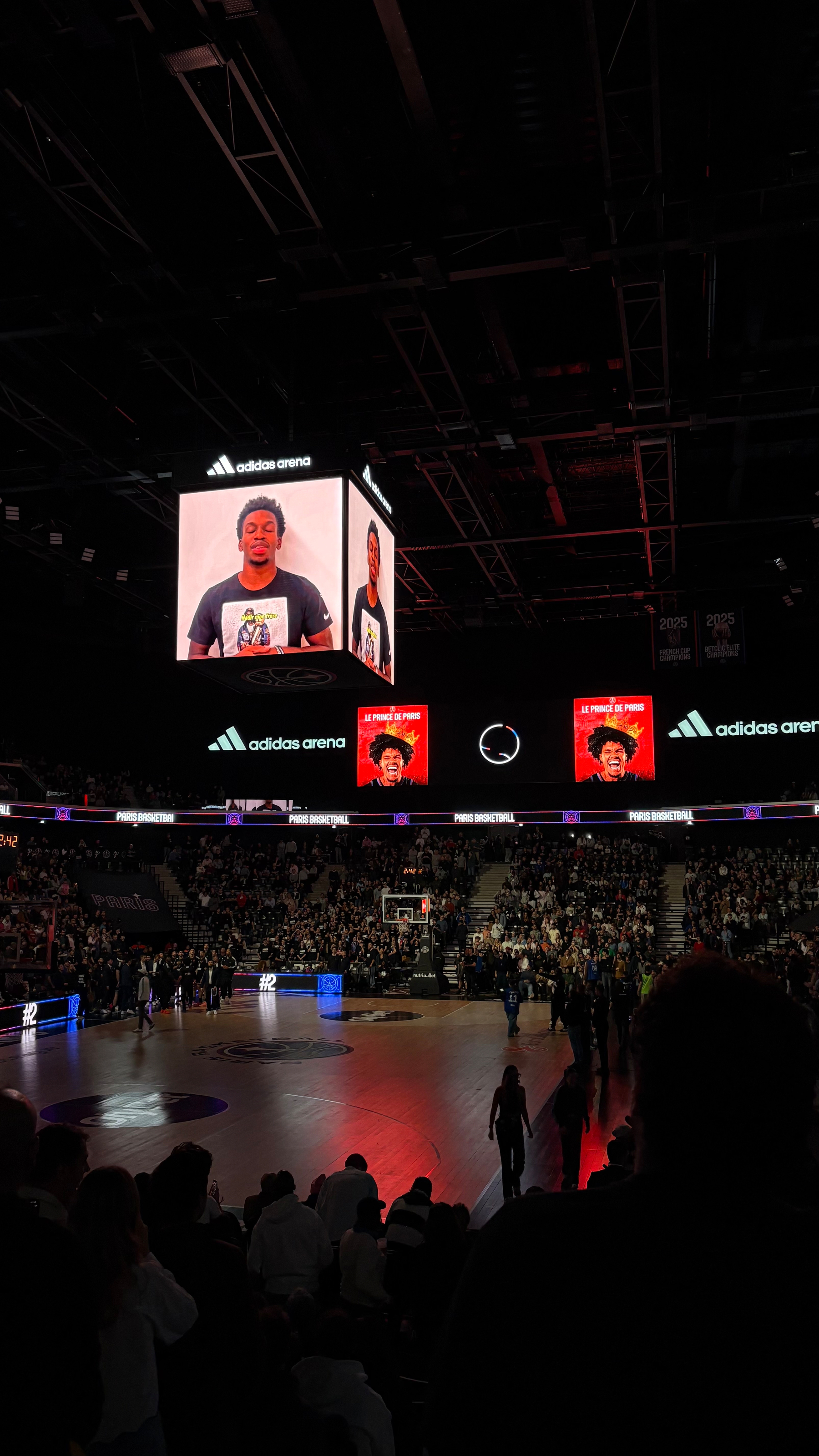 A dimly lit basketball arena with a large central screen displaying a person speaking or presenting, surrounded by an audience and players on the court.