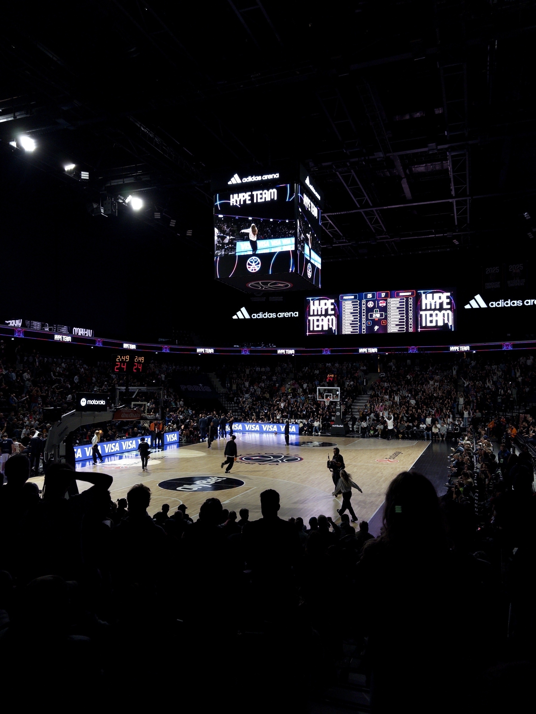 A basketball game is taking place in an arena filled with spectators, with a large screen displaying information overhead.