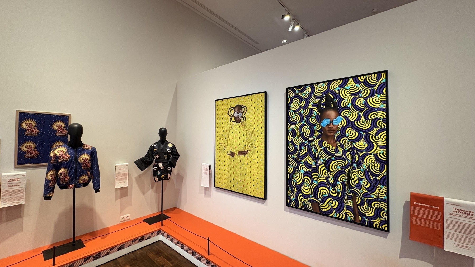 A gallery exhibit showcases vibrant, colorful artworks and patterned clothing on mannequins against a white wall.