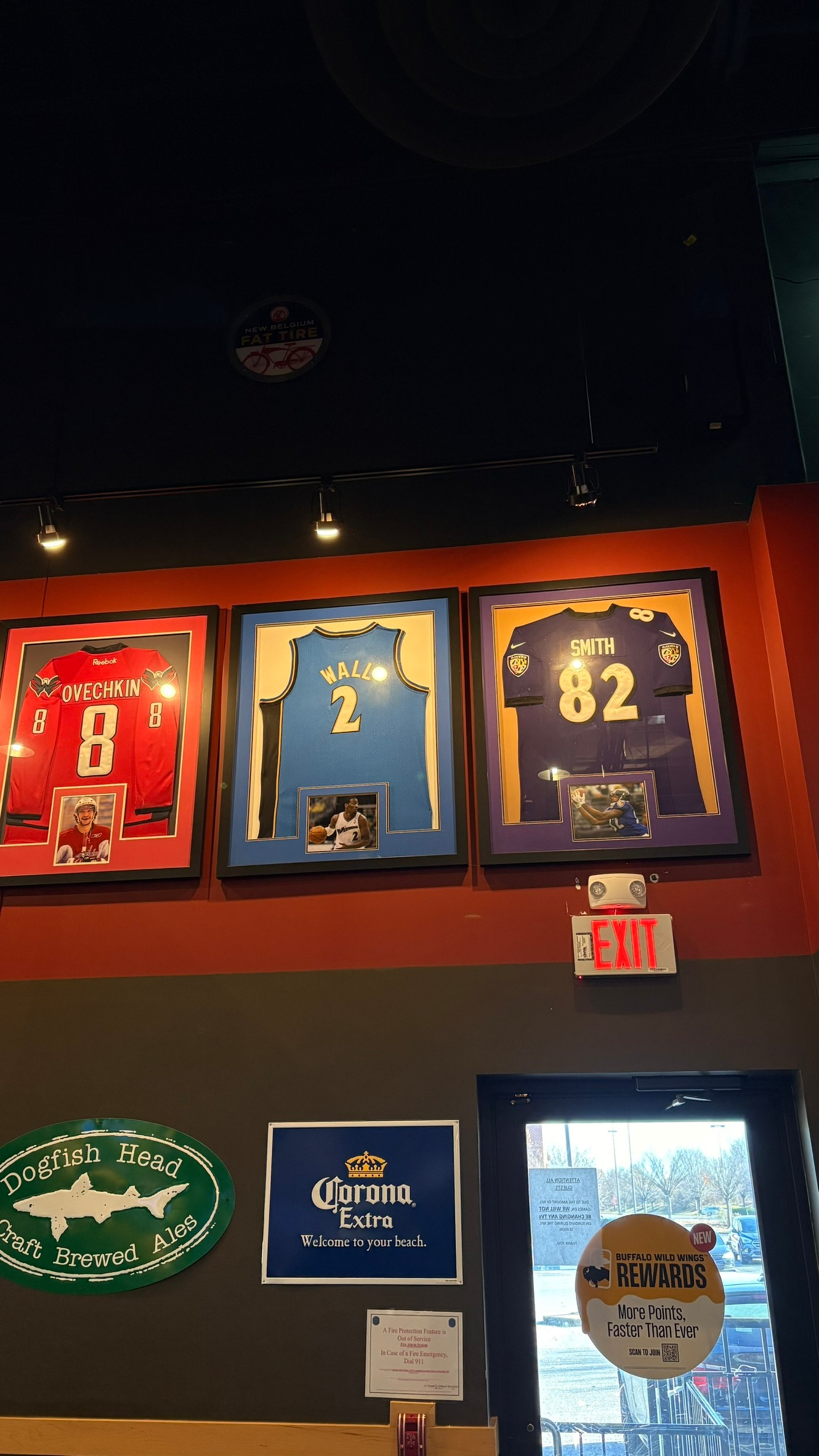 Three framed sports jerseys, one red, one blue, and one purple, are displayed on a wall above a door next to a sign for Corona Extra.