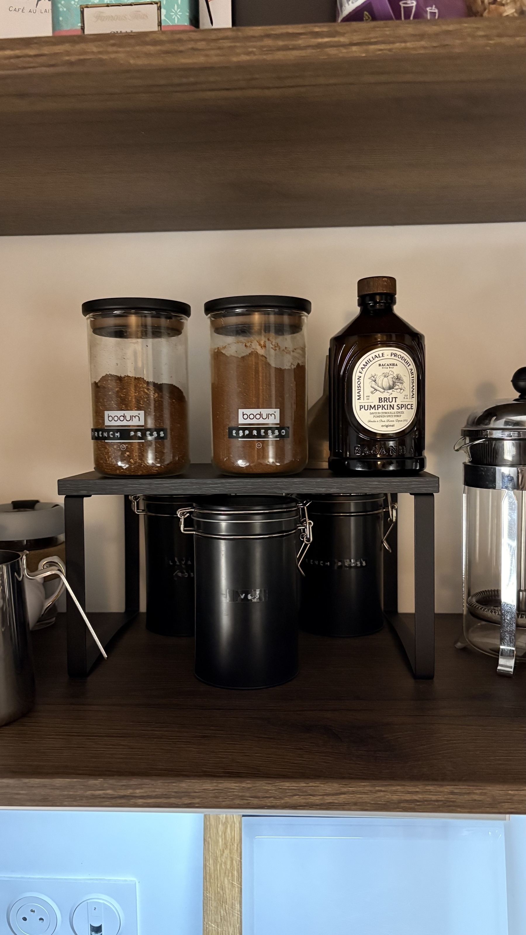 Auto-generated description: A kitchen shelf holds two jars of Bodum coffee, a bottle labeled Maple Pumpkin Spice, and a metal canister beneath a black metal stand.