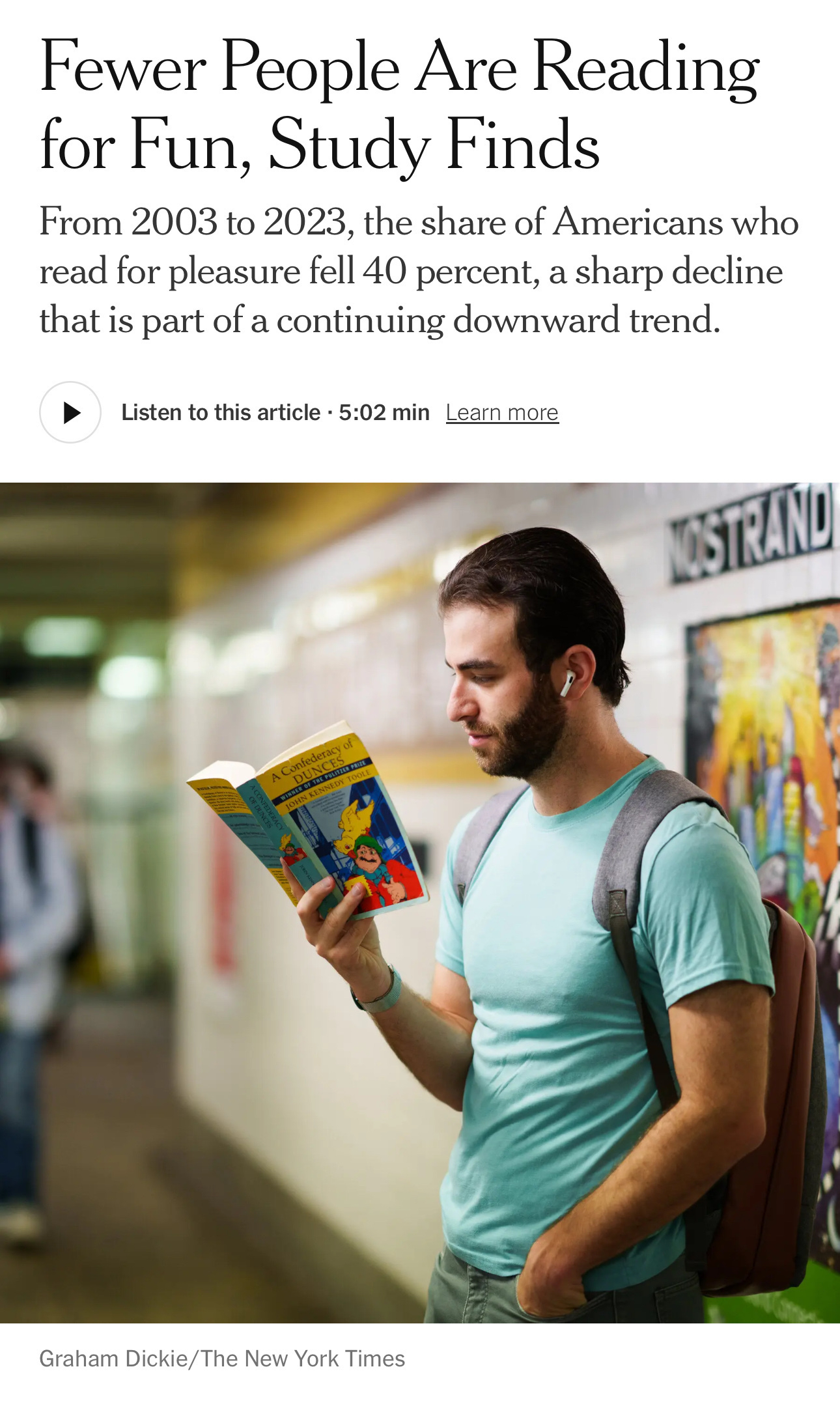 A young man wearing earbuds reads a book while standing in a hallway with blurred people and posters in the background.