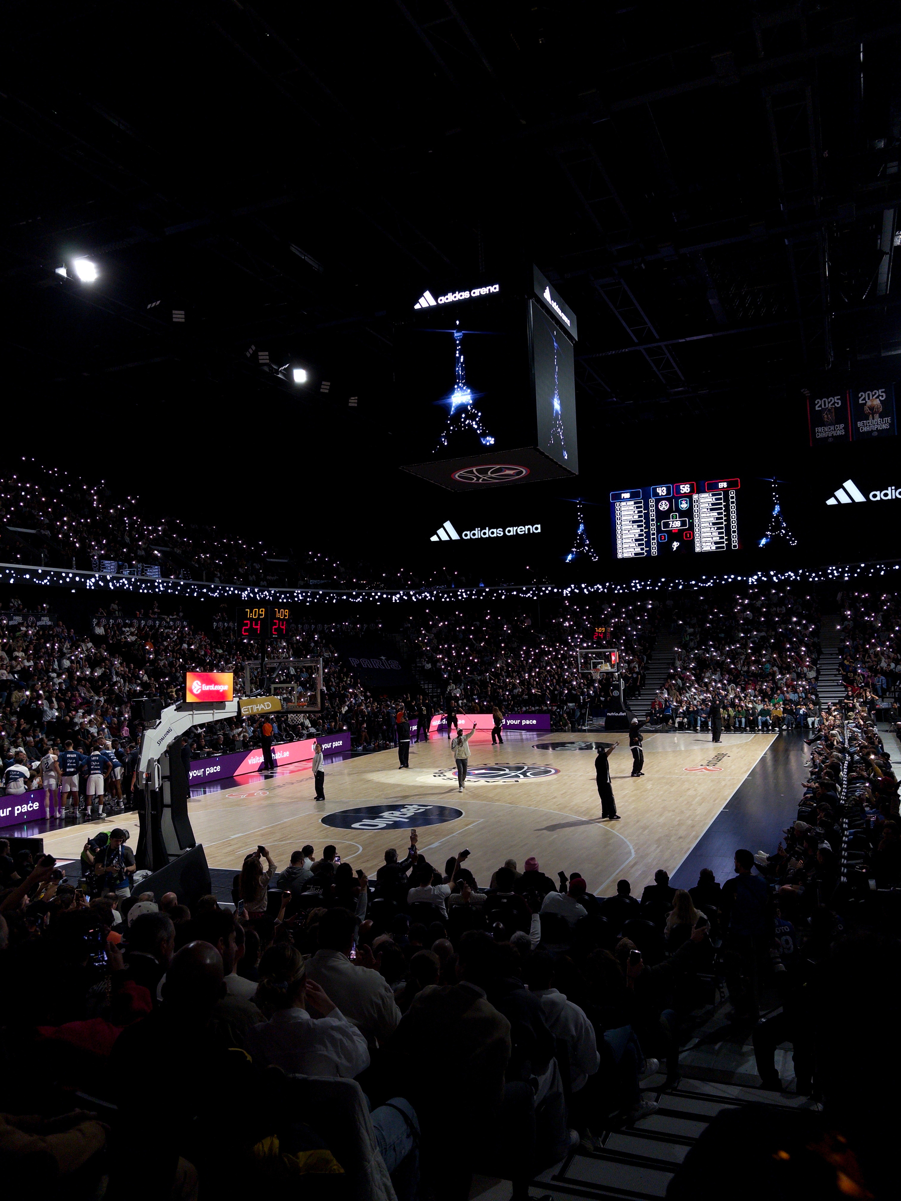 A basketball game is taking place in a packed indoor arena with bright lights and spectators watching the action on the court.