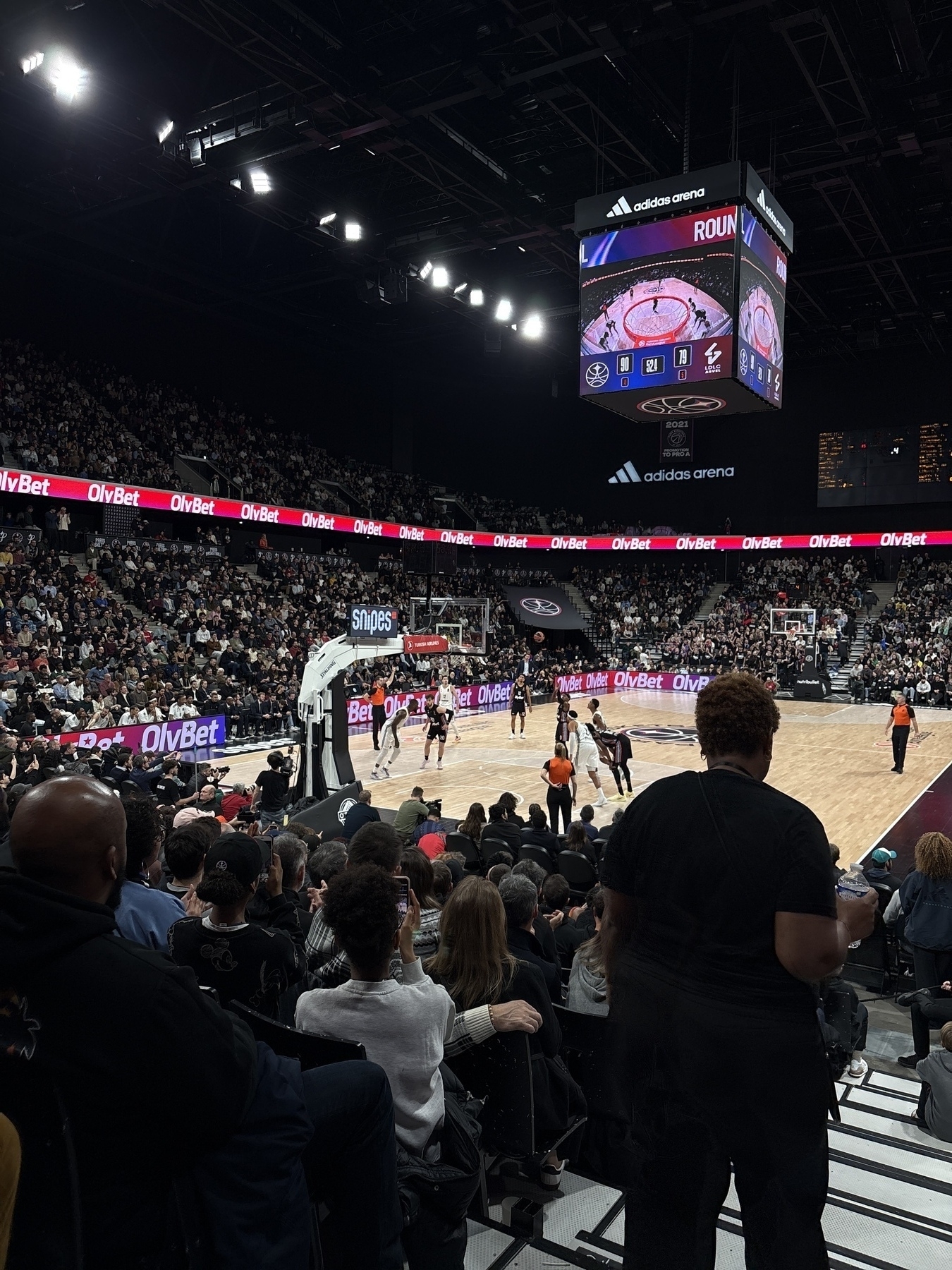 A packed basketball arena features a game in progress with a large screen displaying the action above the court.