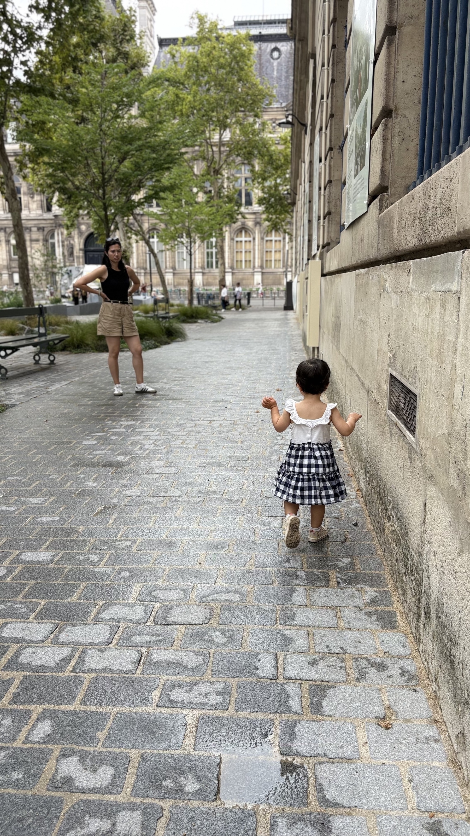 A young child in a checkered dress walks along a cobblestone path with an adult in the background.
