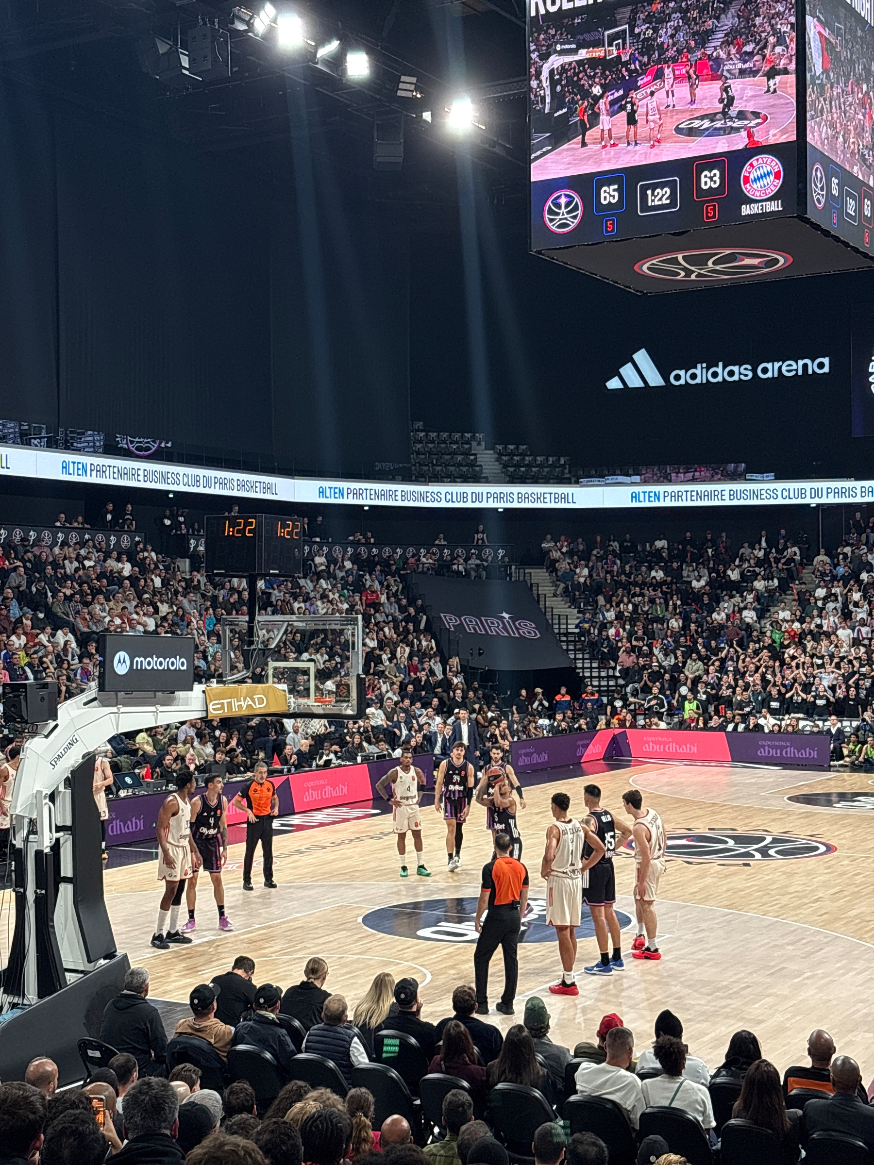 A basketball game is taking place in a large arena with a scoreboard displaying the score and the crowd watching intently.
