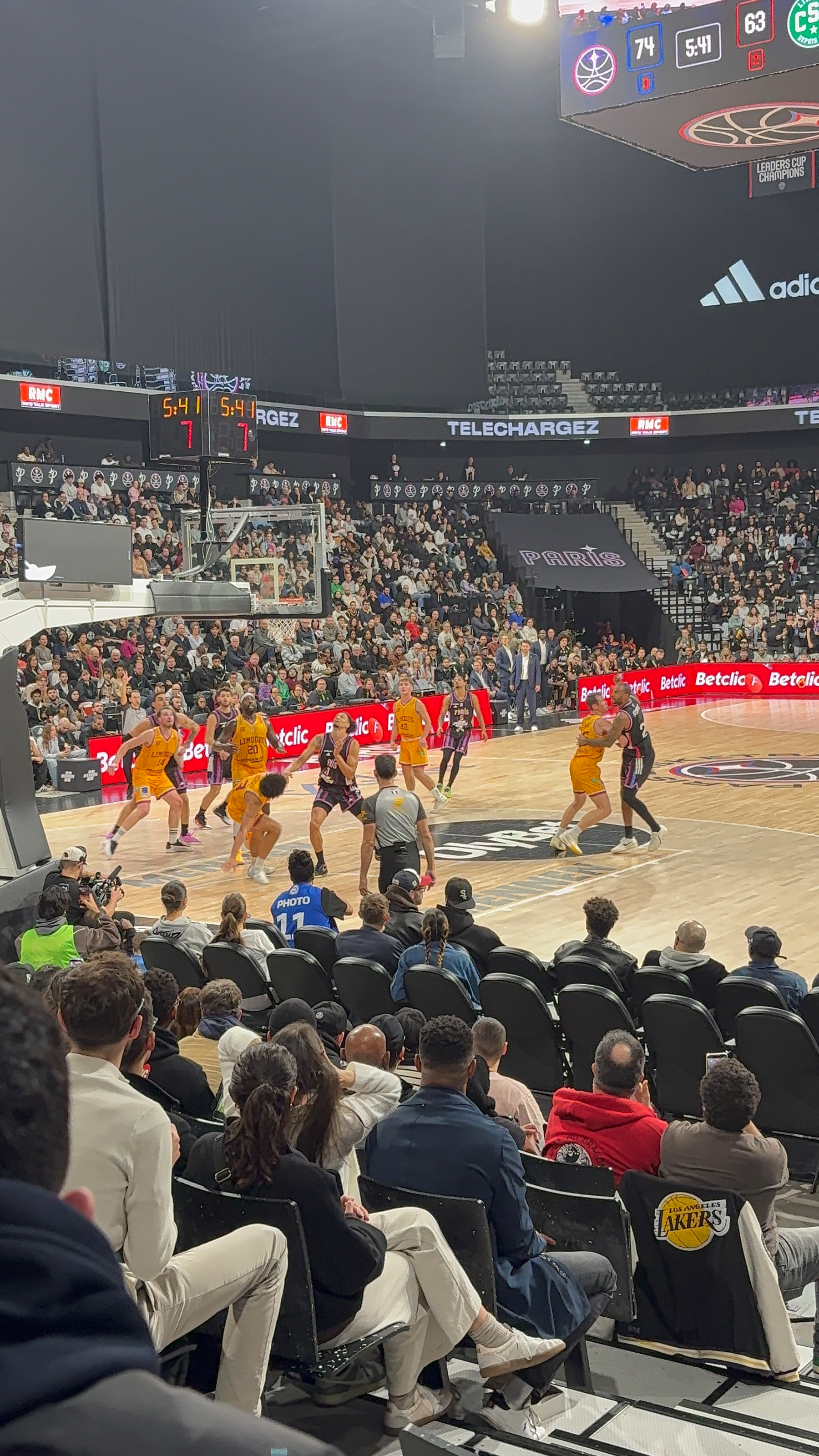 A dance crew performs at center court in an indoor basketball arena with a large screen displaying their performance above.