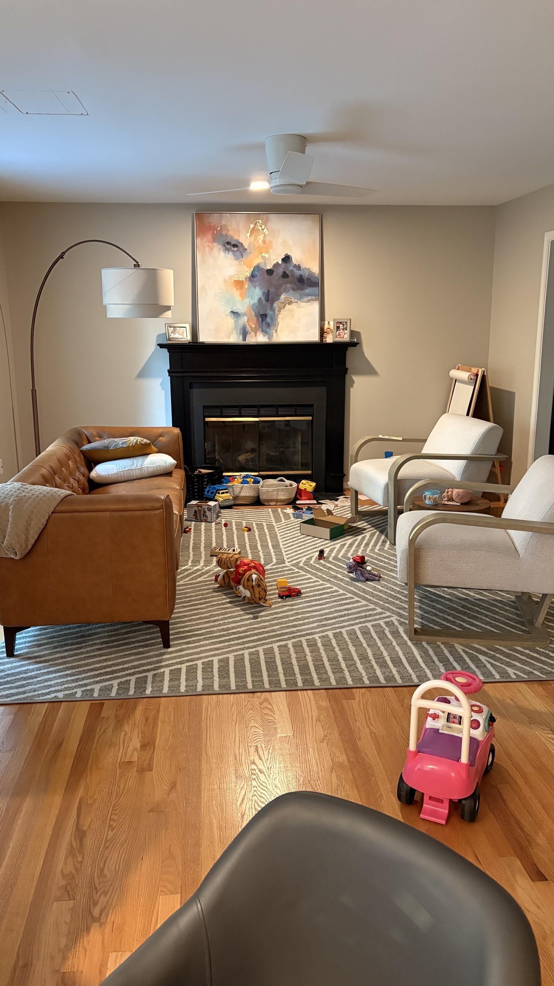 A cozy living room features a fireplace with toys scattered across a patterned rug, surrounded by a leather couch and armchairs.