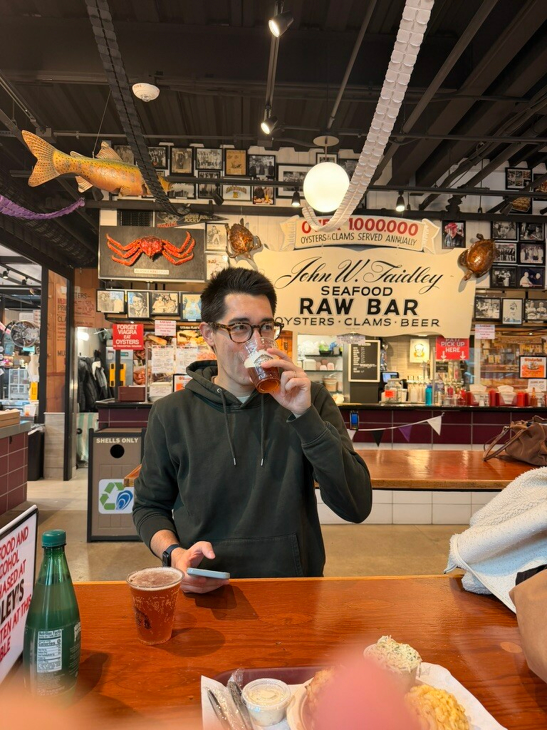 A man in a casual setting drinks from a plastic cup in a seafood restaurant with various decorations and a sign about oysters in the background.