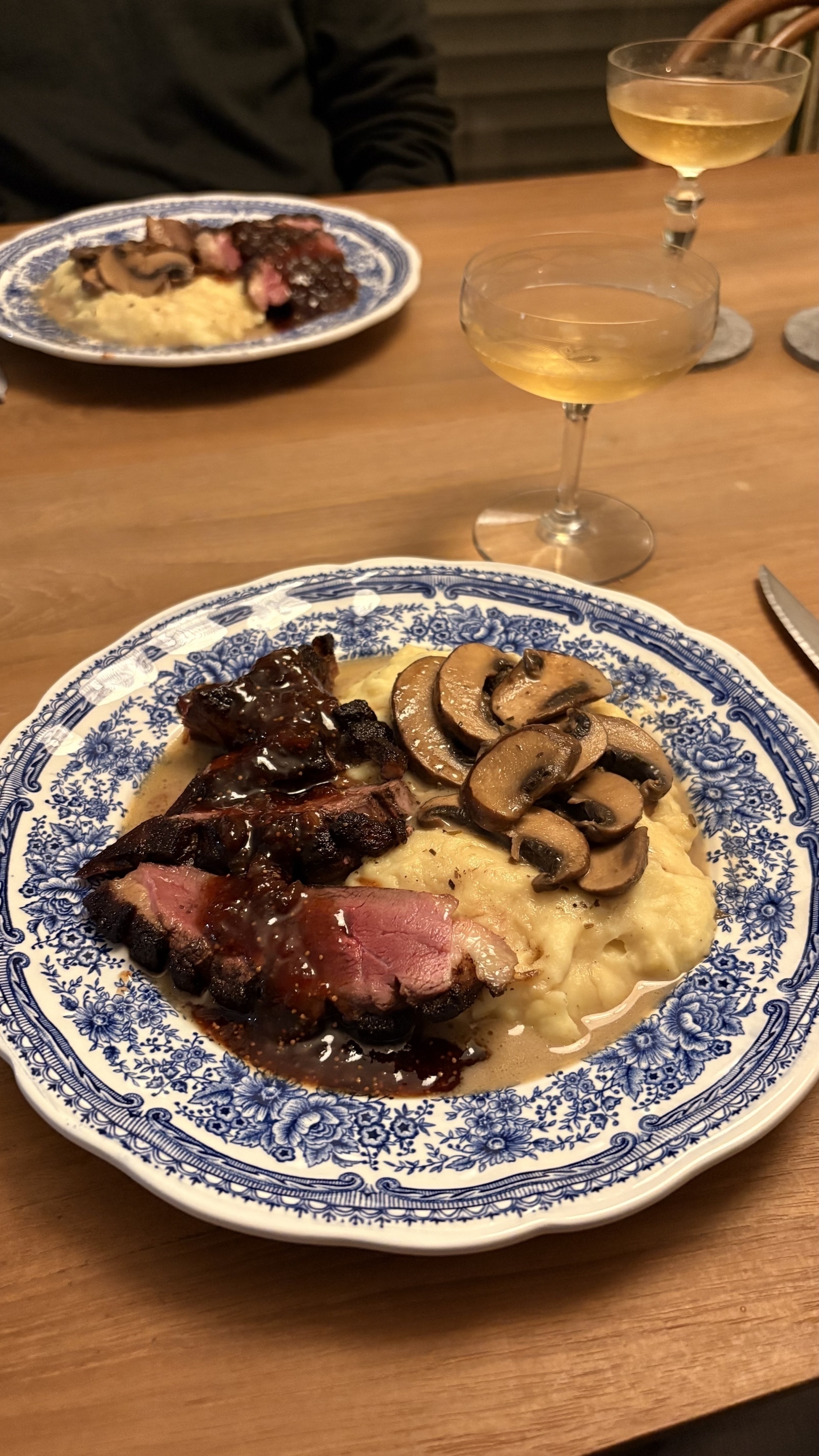 A dinner plate features sliced meat with a dark sauce, mushrooms, and mashed potatoes, accompanied by a glass of white wine, set on a wooden table.