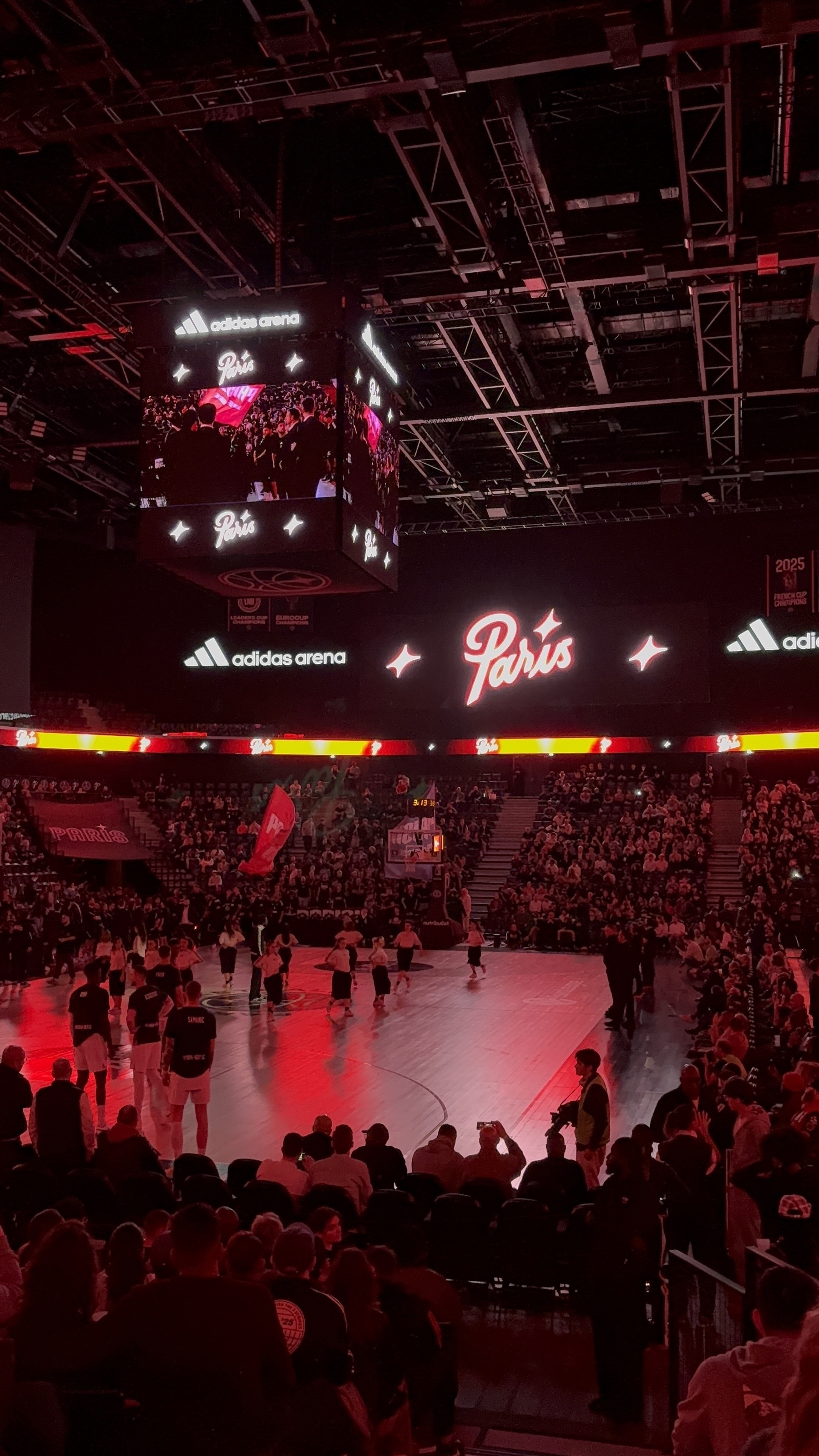 A sports arena is lit up in red lights with a Paris sign on the screen and a group of people on the court.