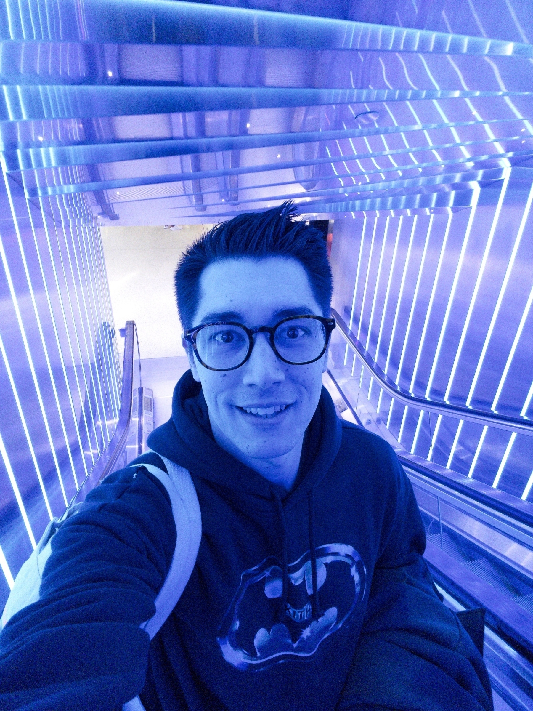 A person wearing glasses and a Batman hoodie takes a selfie in a blue-lit environment with an escalator in the background.