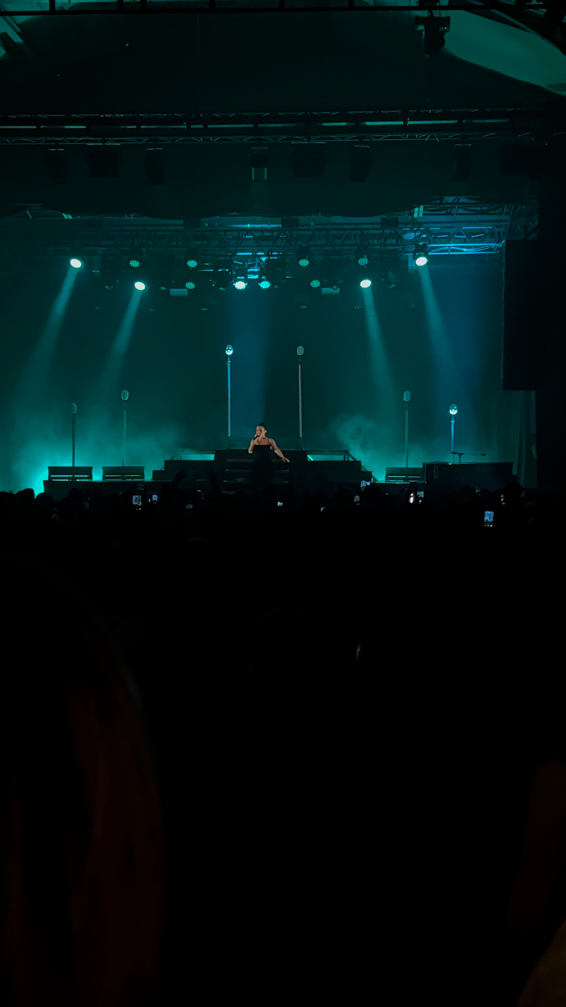 A performer is on stage under teal lighting, with an audience in the dark foreground.