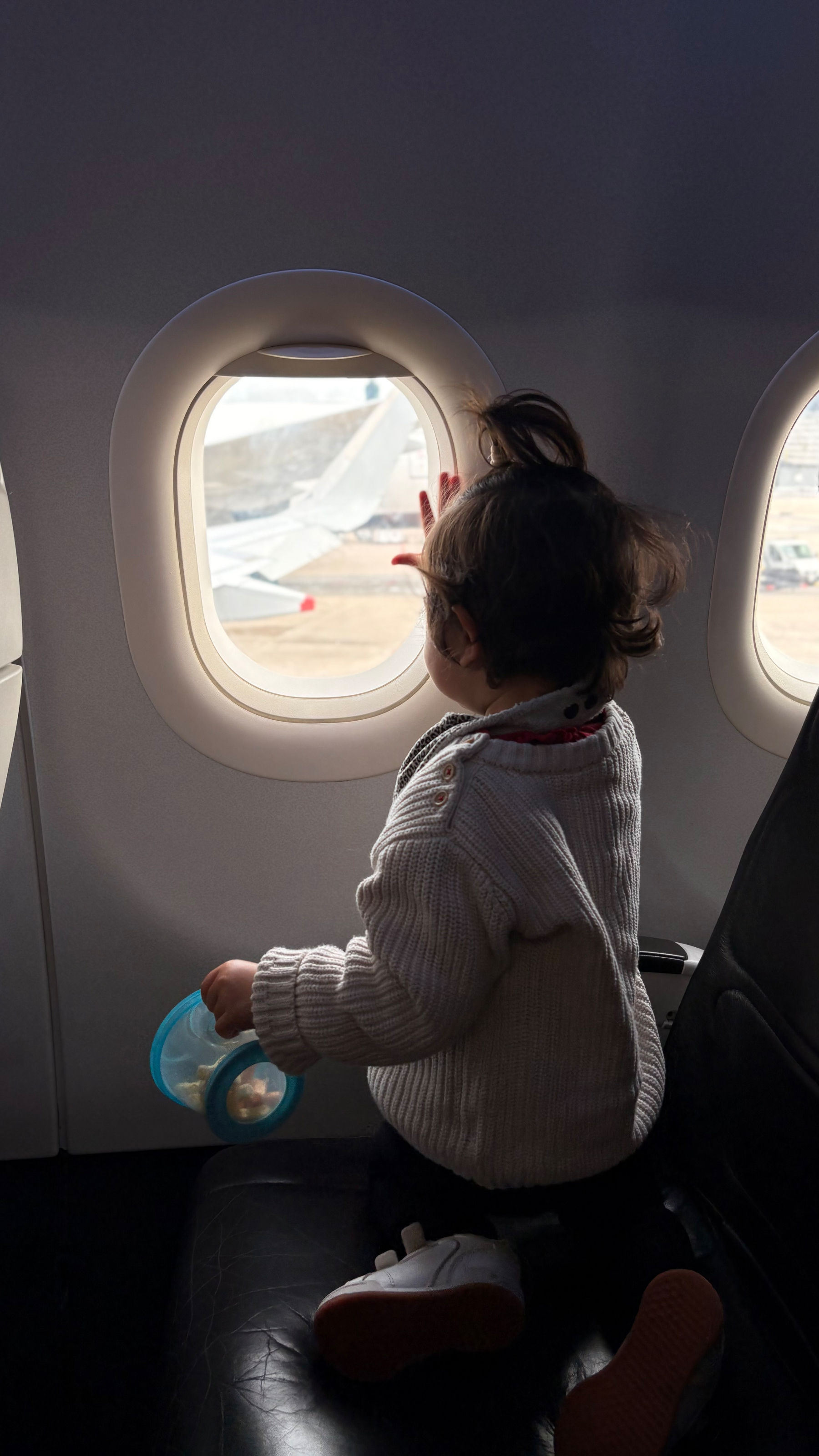A small child sits on an airplane seat, looking out the window while holding a container.