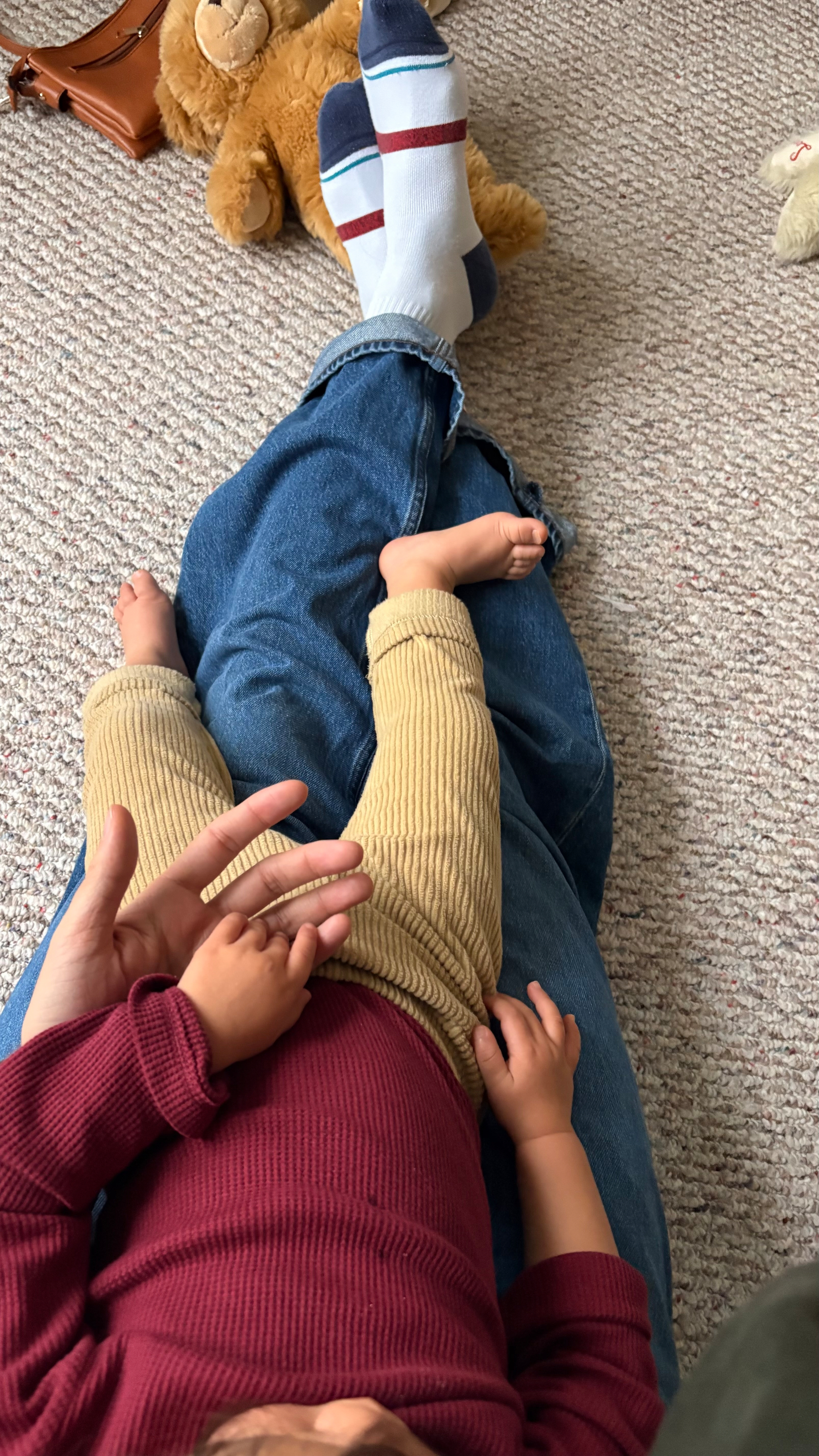 A child wearing a maroon top and beige pants is lying on a person's lap, with striped socks and a teddy bear in the background.