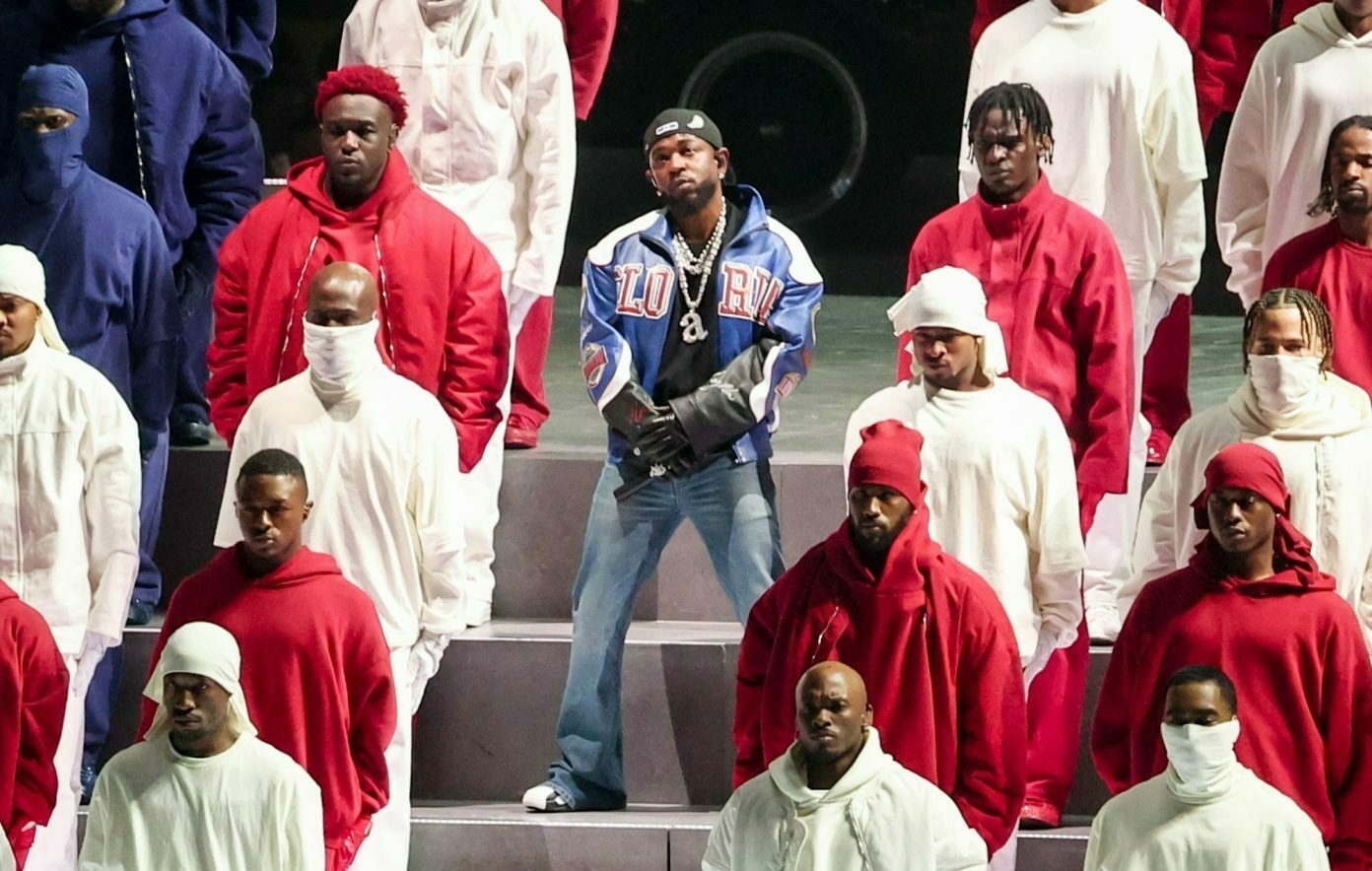 A group of performers wearing red and white outfits surrounds a central figure dressed in a blue jacket with LUXURY written on it, set on a stage.