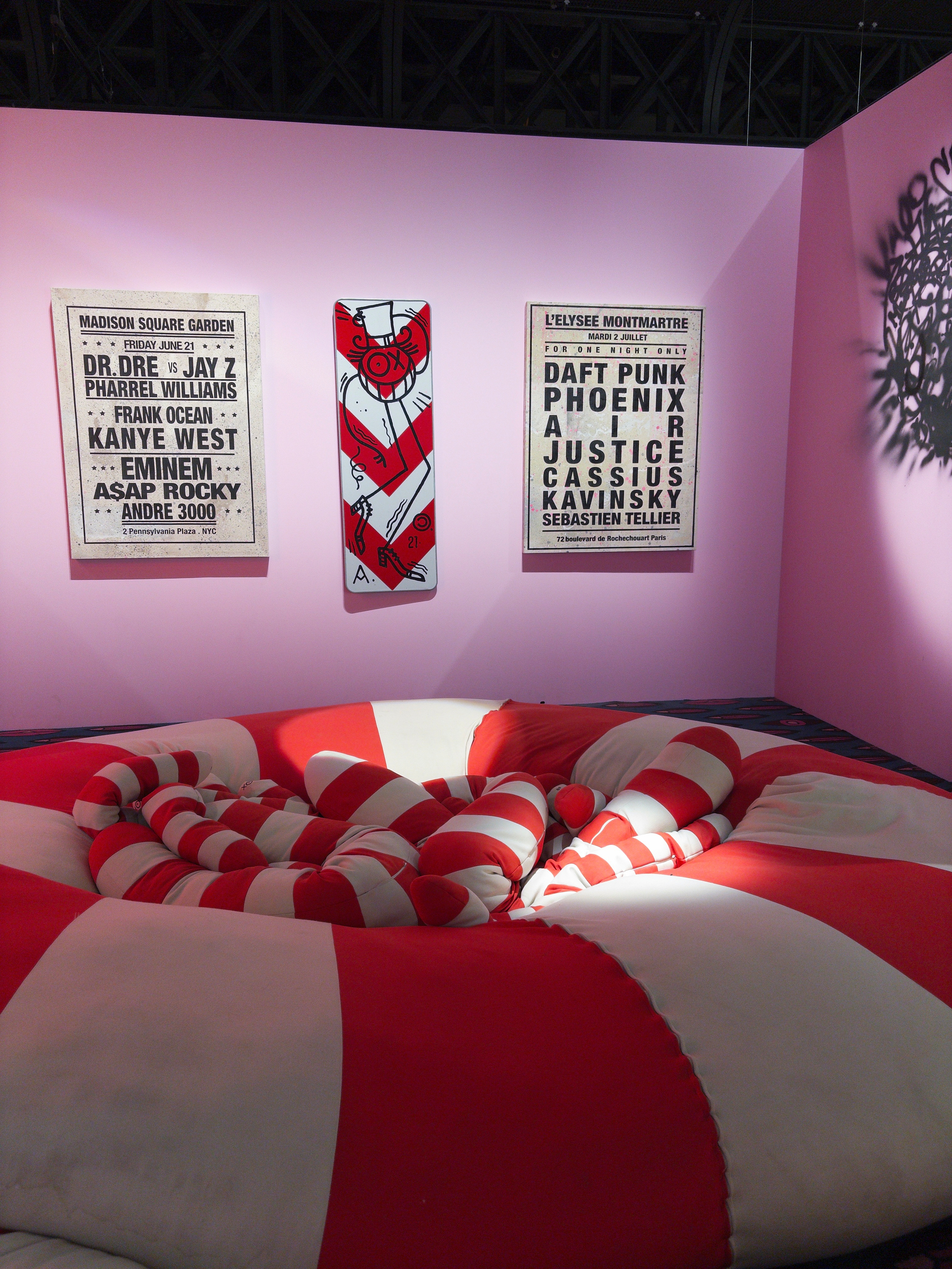 A cozy, oversized cushioned seat with red and white stripes is set against a pink wall adorned with music event posters and a skateboard deck featuring a red and white design.