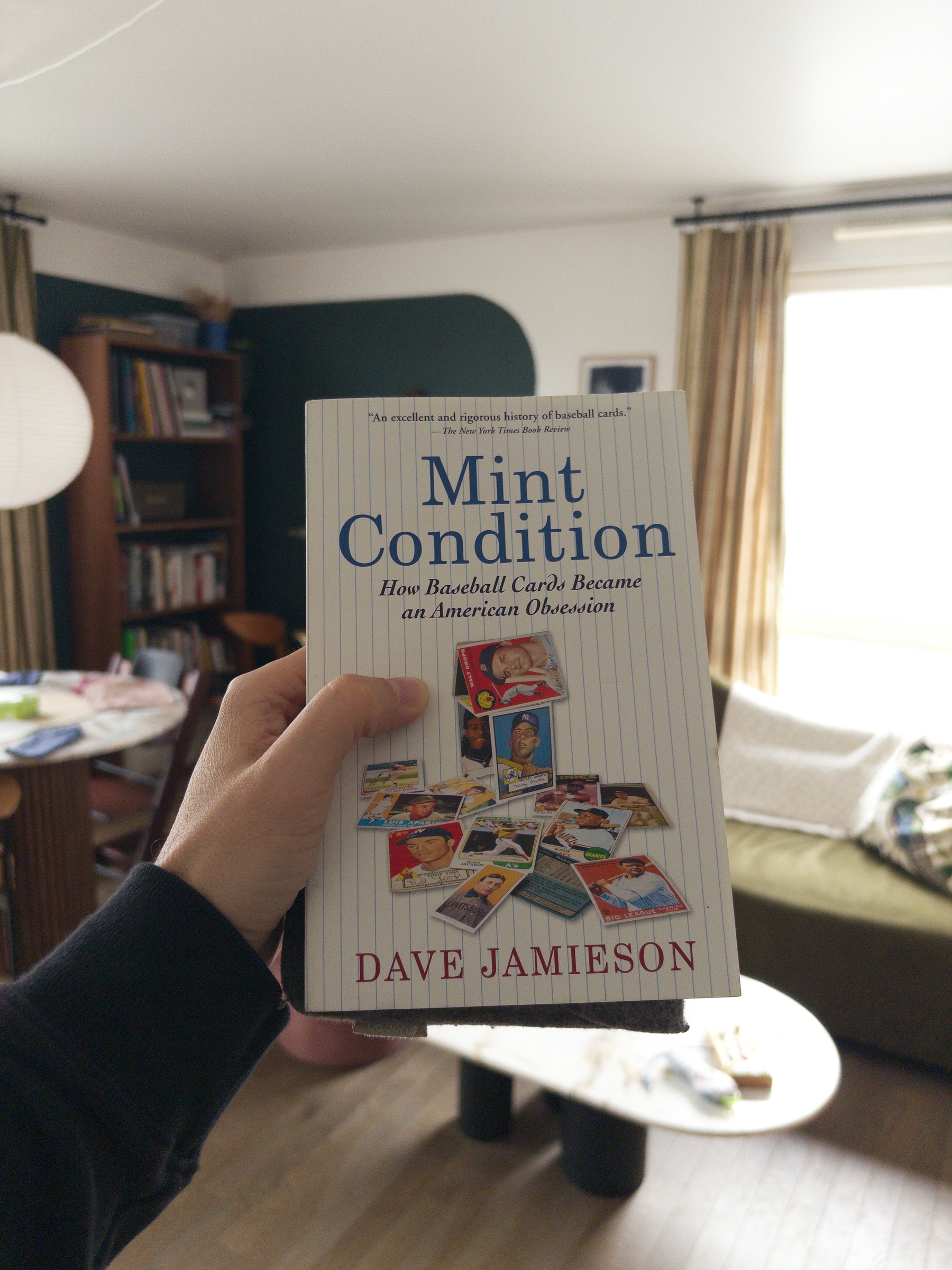 A hand is holding a book titled Mint Condition: How Baseball Cards Became an American Obsession by Dave Jamieson in a living room.