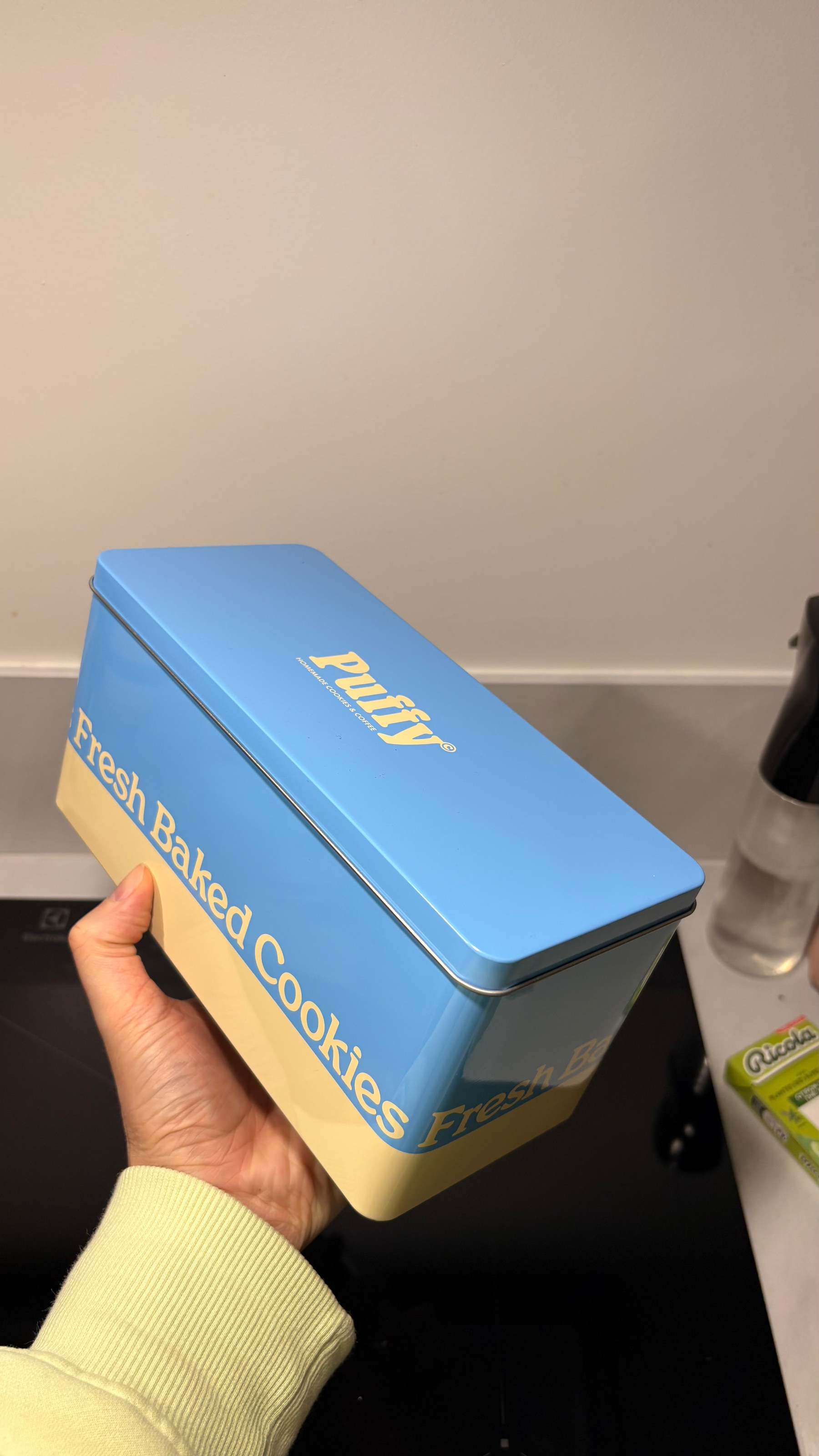 A person is holding a blue and cream-colored cookie tin labeled Fresh Baked Cookies with the brand Puffy on top.