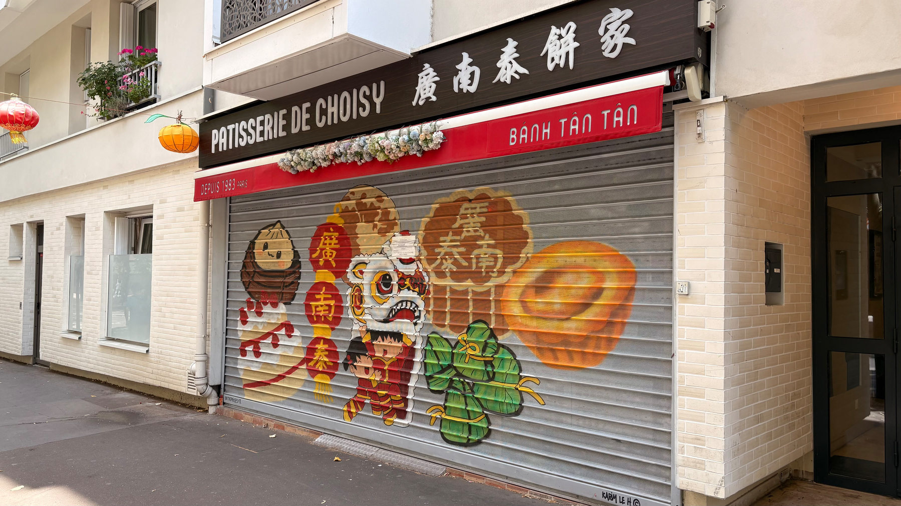 A storefront with a shutter painted to depict various traditional Asian pastries, a lion dance head, and other festive decorations.
