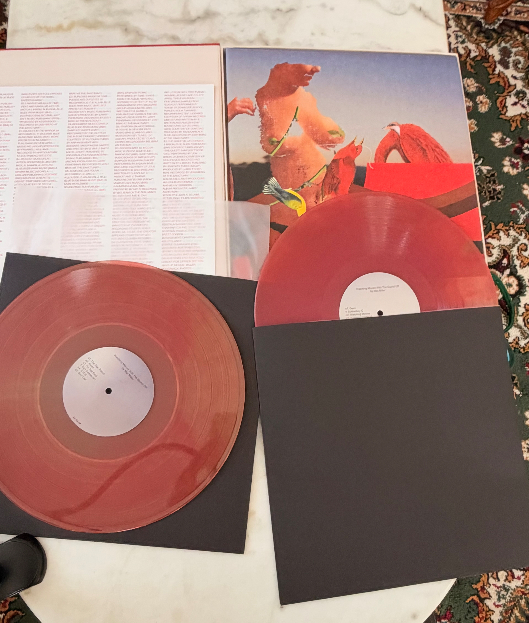 Auto-generated description: Two translucent red vinyl records are displayed alongside an album cover featuring surreal artwork of a topless figure with birds, with one vinyl partially out of its black sleeve and the other fully visible.