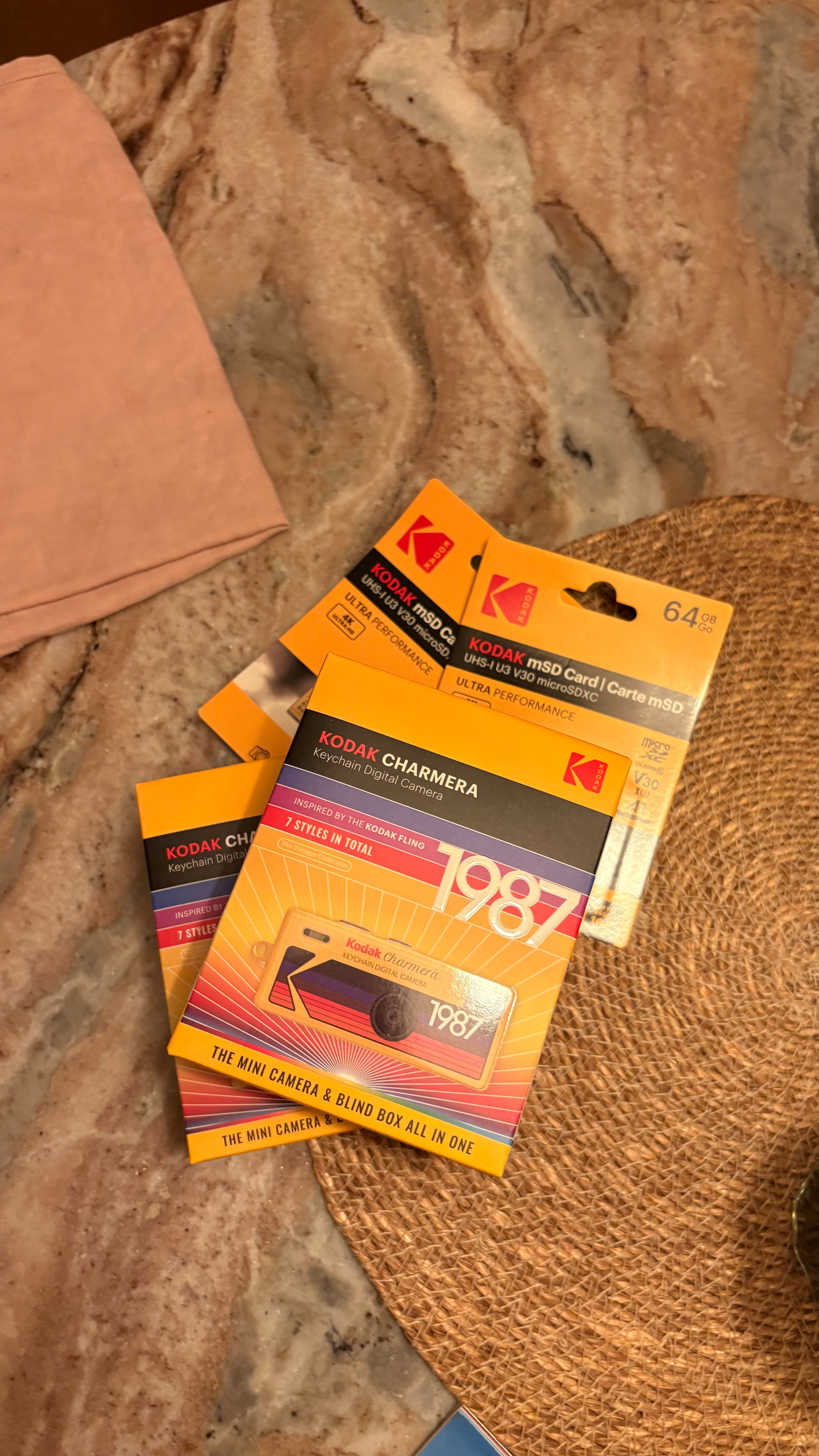 Several Kodak CharmEra keychain digital cameras and Kodak 64 GB microSD cards are placed on a marble surface next to a woven mat and a folded fabric.