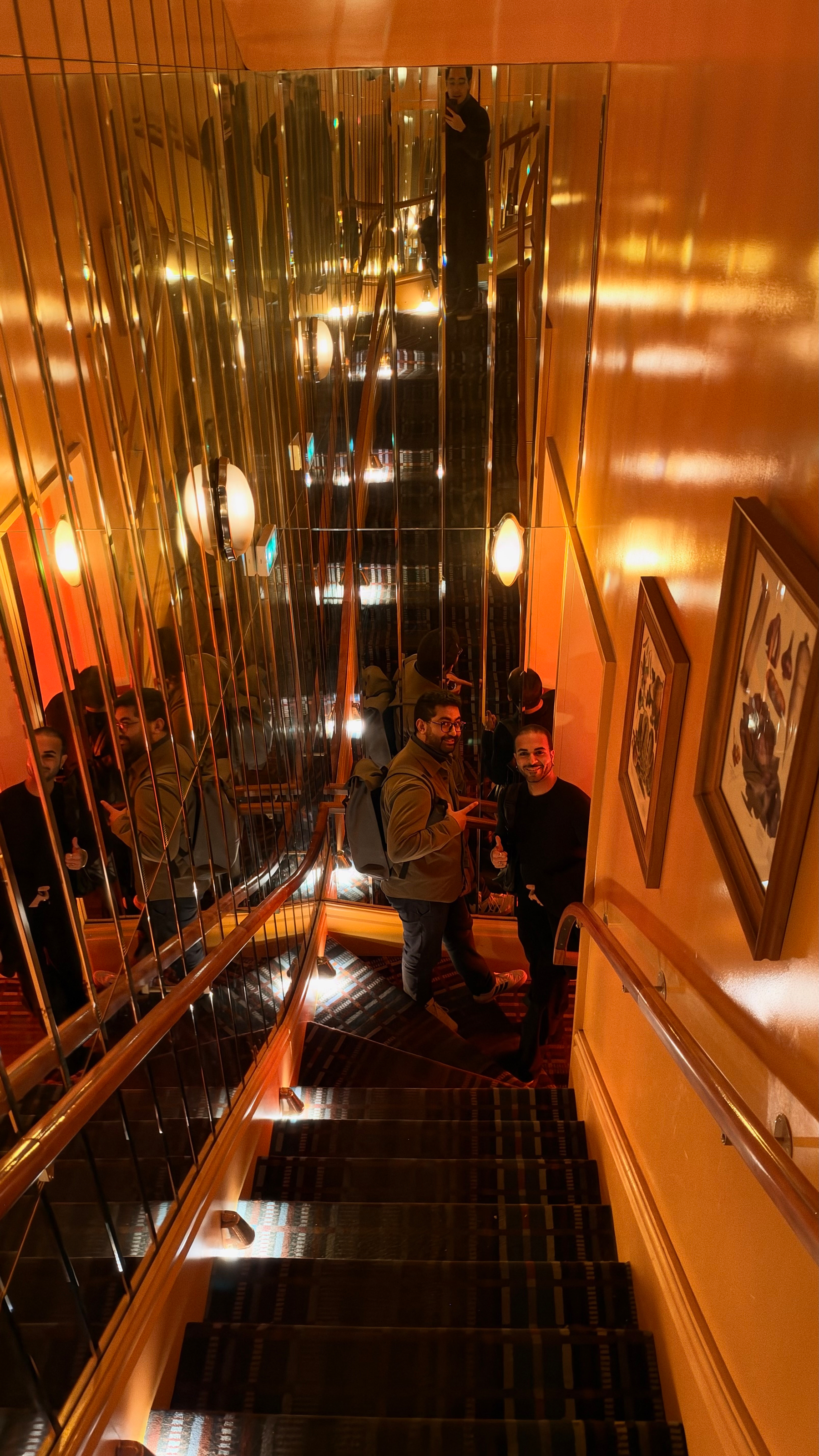 Two men are standing and posing on a carpeted staircase with mirrored walls reflecting their images and warm lighting illuminating the scene.