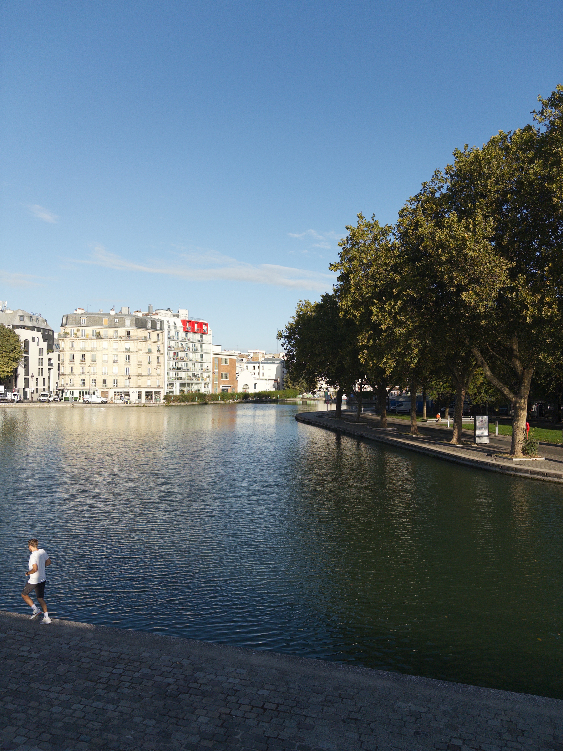 A calm river or canal flows through a cityscape with buildings on one side and a tree-lined walkway on the other, while a person walks along the water's edge.