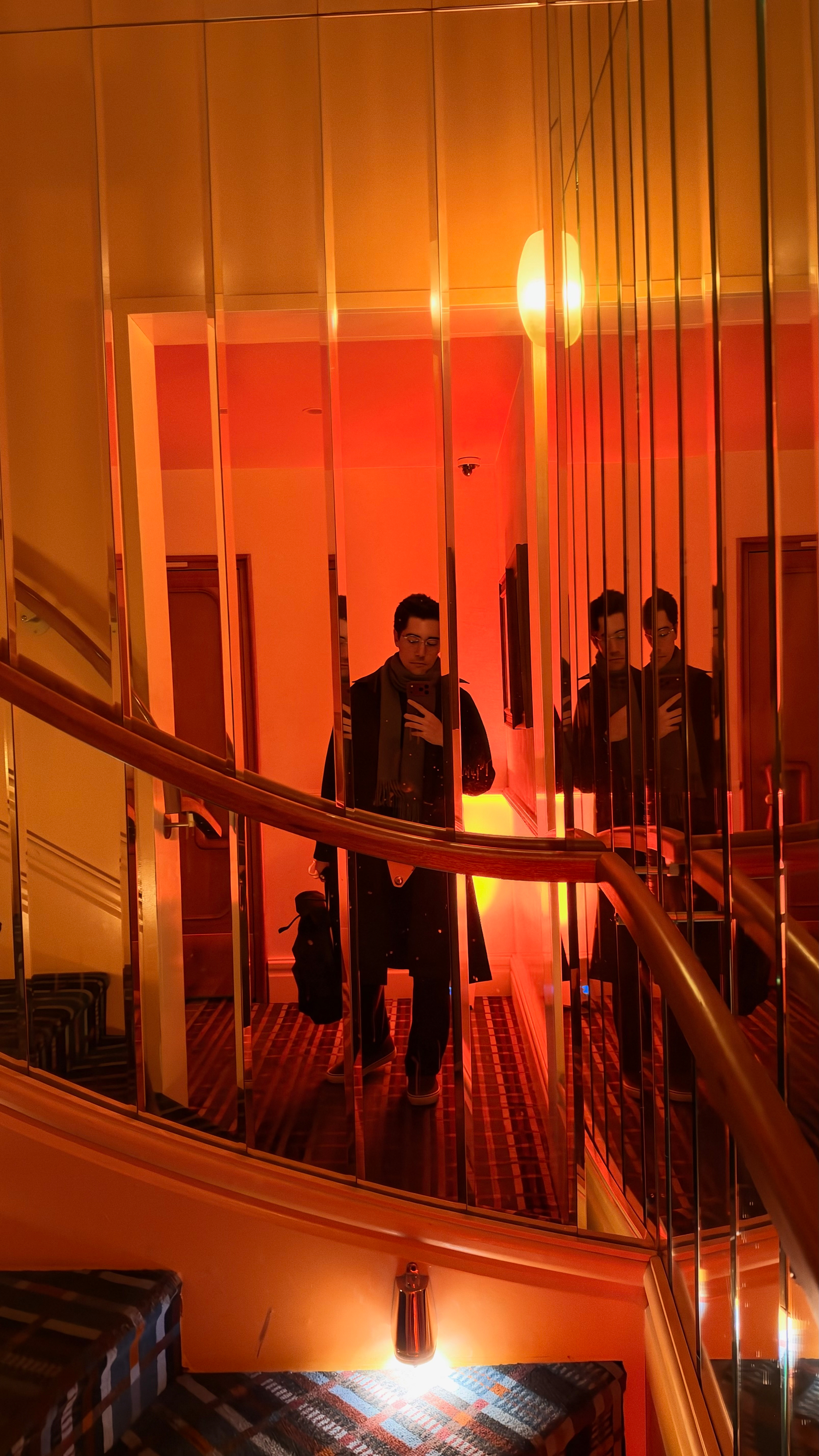 A person is taking a mirror selfie on a staircase with warm orange lighting and multiple reflections in vertical mirrored panels.