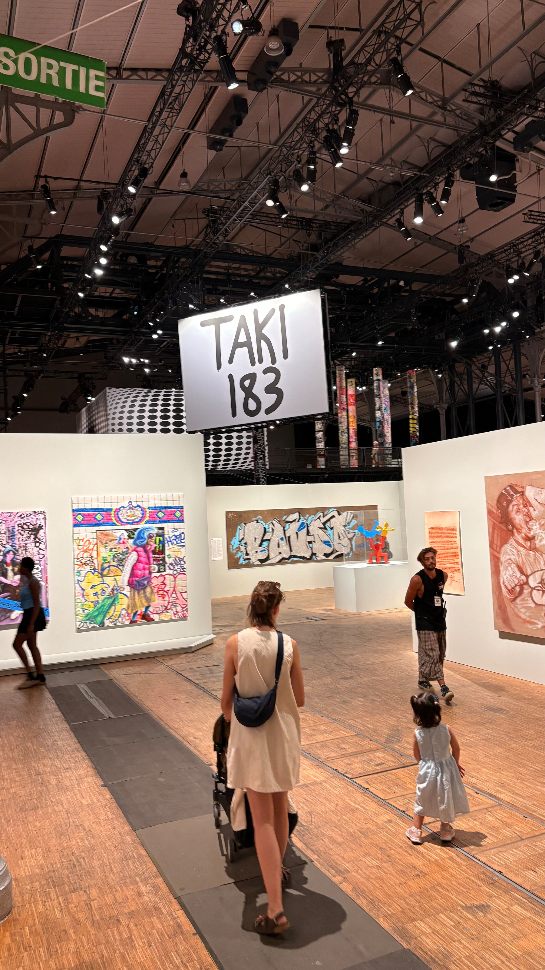 A gallery space features various colorful artworks on the walls and people, including a woman pushing a stroller and a child walking alongside her, exploring the exhibits under the sign TAKI 183.