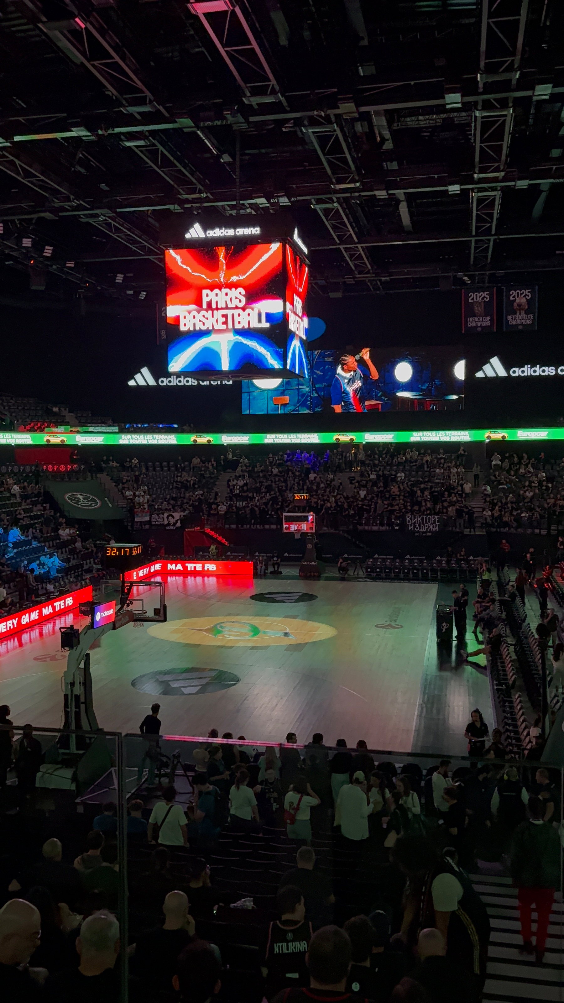 A basketball arena is filled with spectators and illuminated by a large scoreboard displaying Paris Basketball under the Adidas Arena branding.