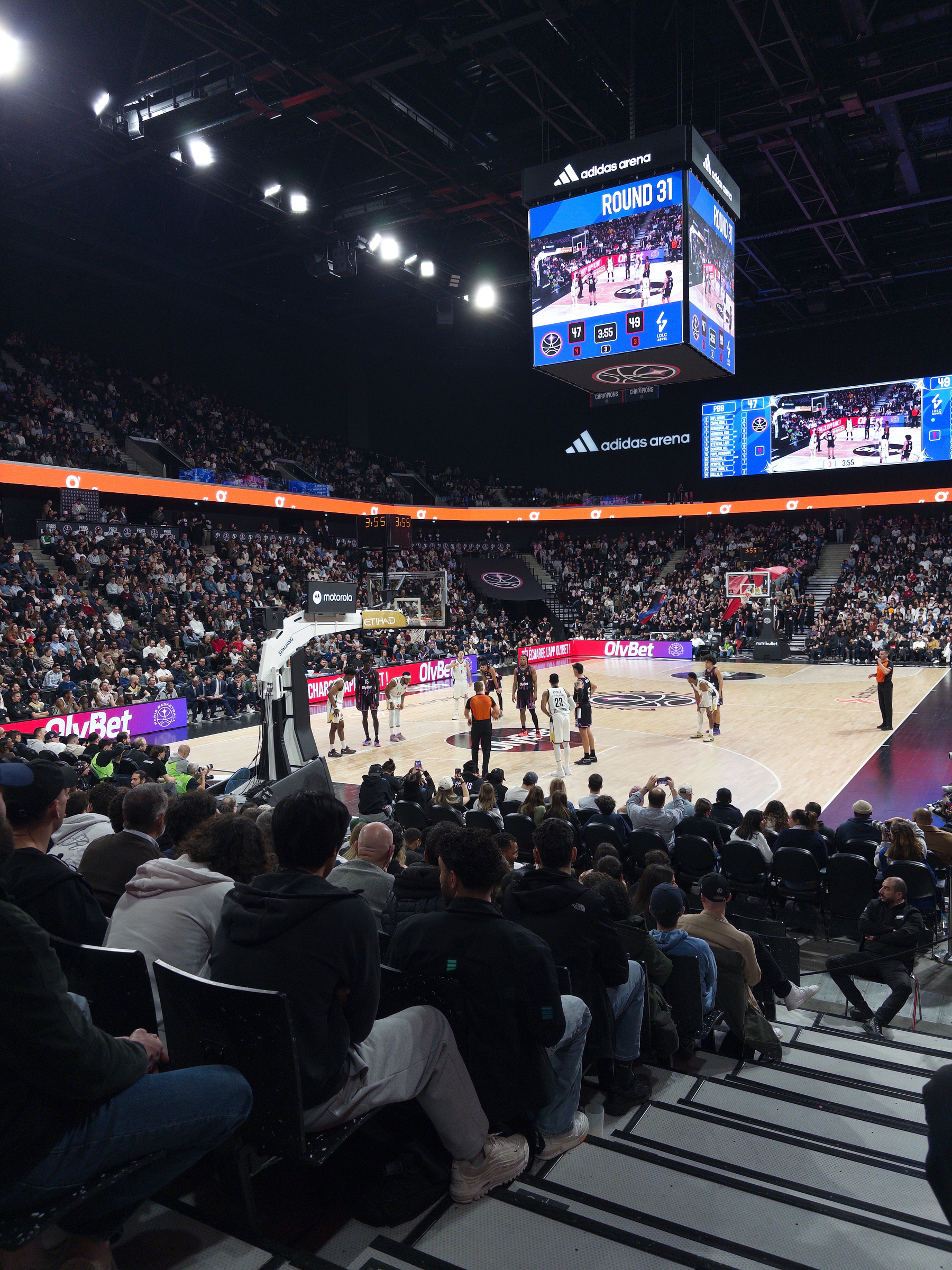 A basketball game is taking place in a packed arena with spectators watching from the stands.
