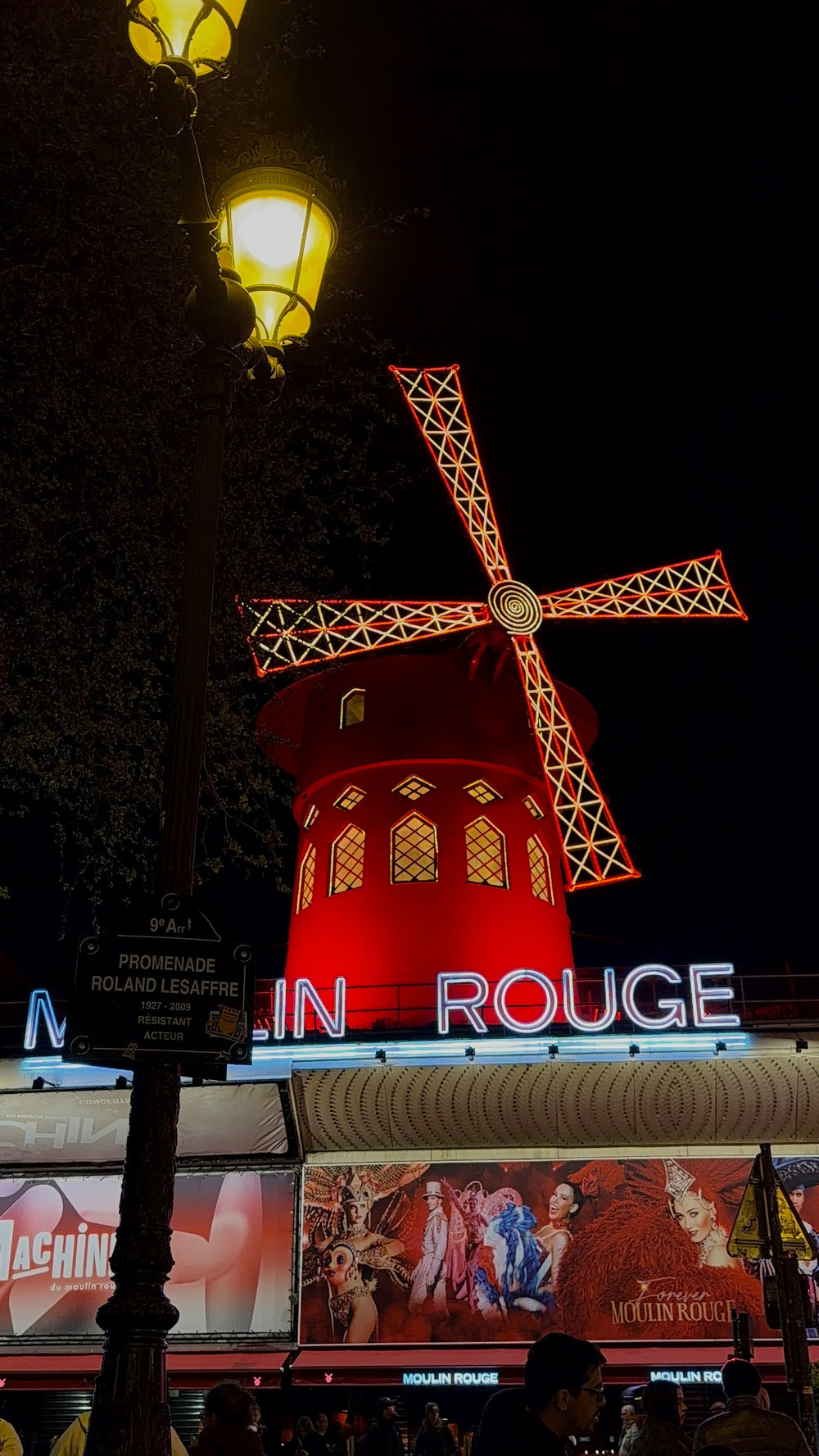 A red windmill with illuminated sails and the words Moulin Rouge in bright neon are showcased against a nighttime backdrop.