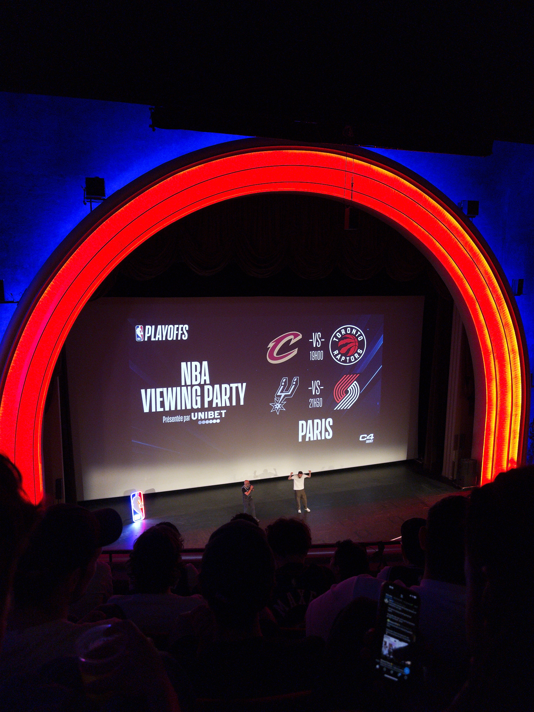 An NBA viewing party is taking place inside a theater with a large screen displaying matchups, featuring a vibrant red arched lighting design.