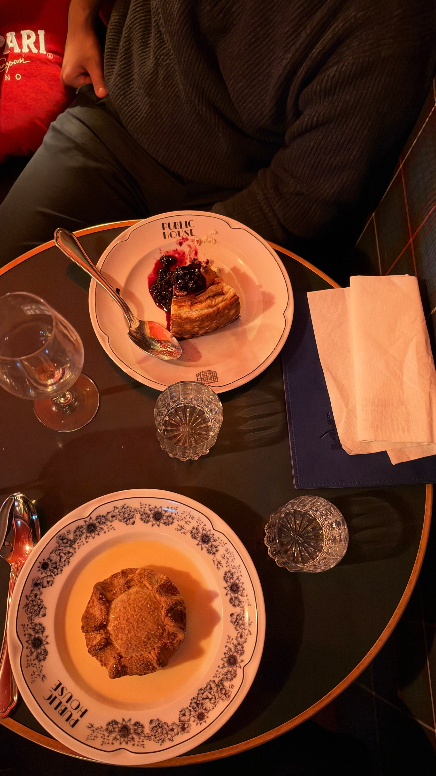 Two plates with pastries and jam, two glasses, a spoon, a fork, and a napkin are arranged on a round table next to a seated person.
