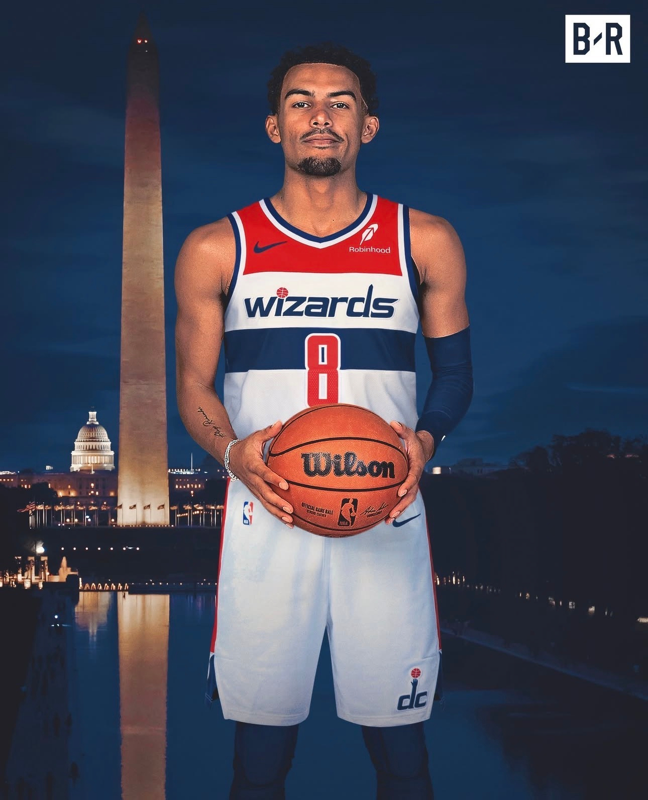 A basketball player wearing a Washington Wizards uniform stands holding a basketball with the Washington Monument in the background.