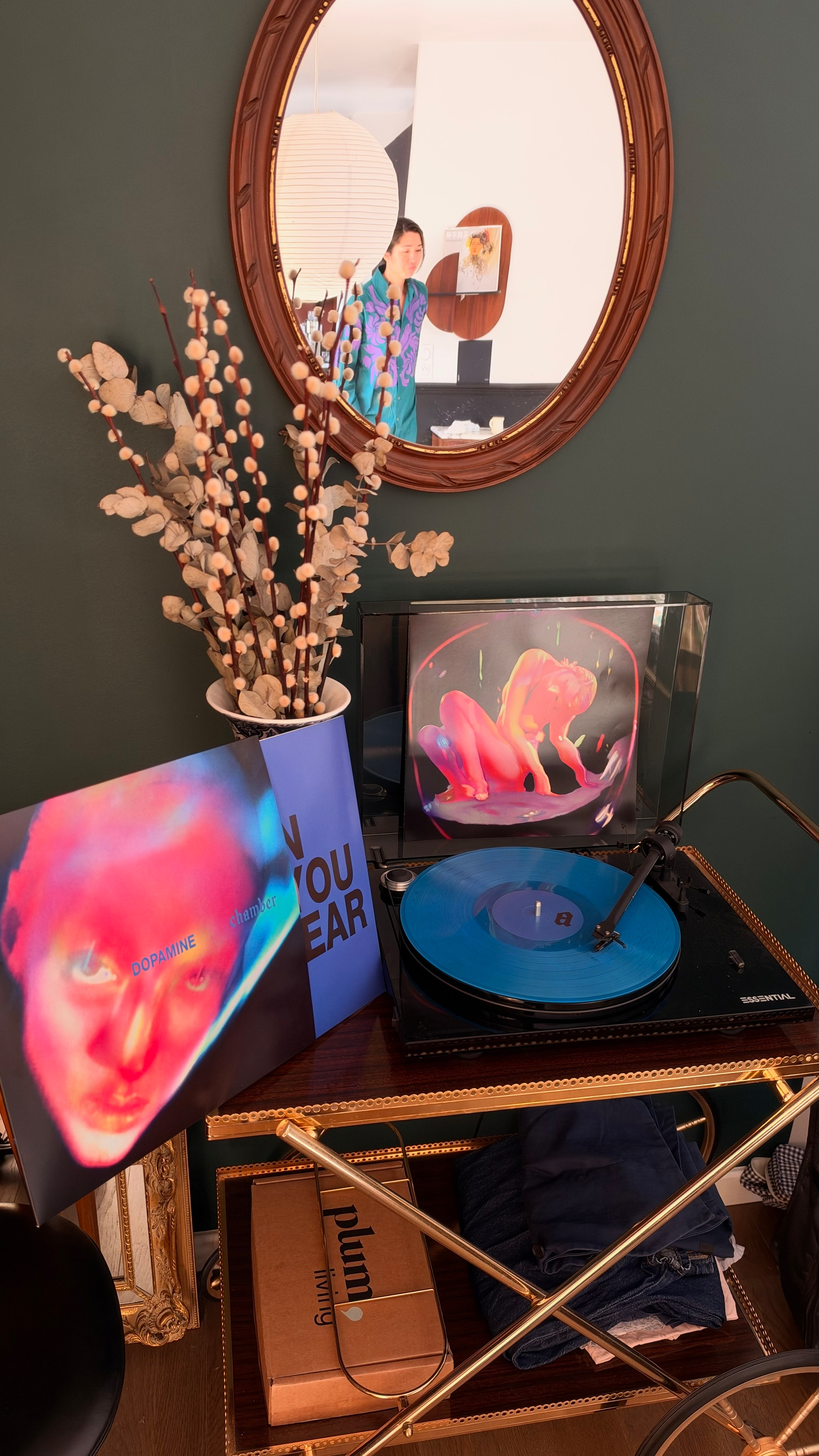 A vintage turntable playing a blue vinyl record is set on a cart with album covers, a vase with dried flowers, and a round mirror reflecting a person standing in the background.