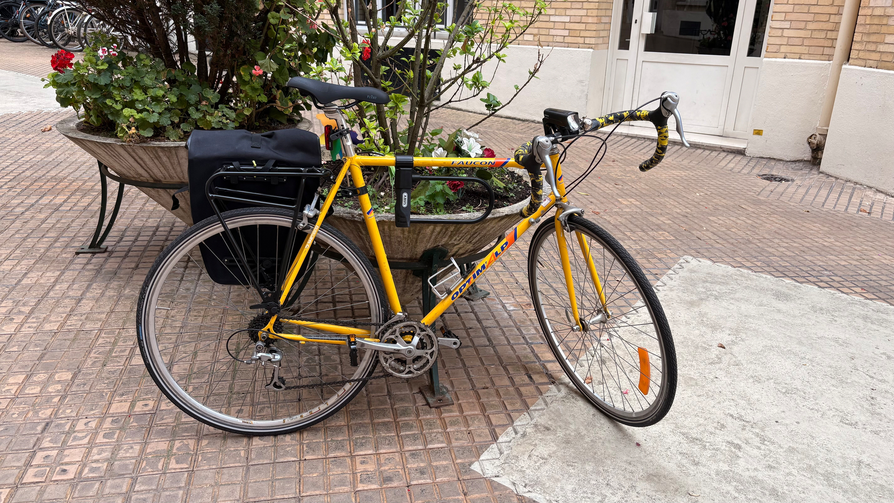 A yellow road bicycle with saddlebags is parked next to a bench with plants on a brick-paved sidewalk.