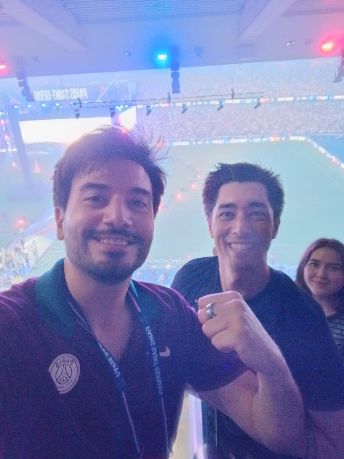 Two people are smiling and posing for a selfie in a stadium with a sports field in the background, along with another person standing behind them.
