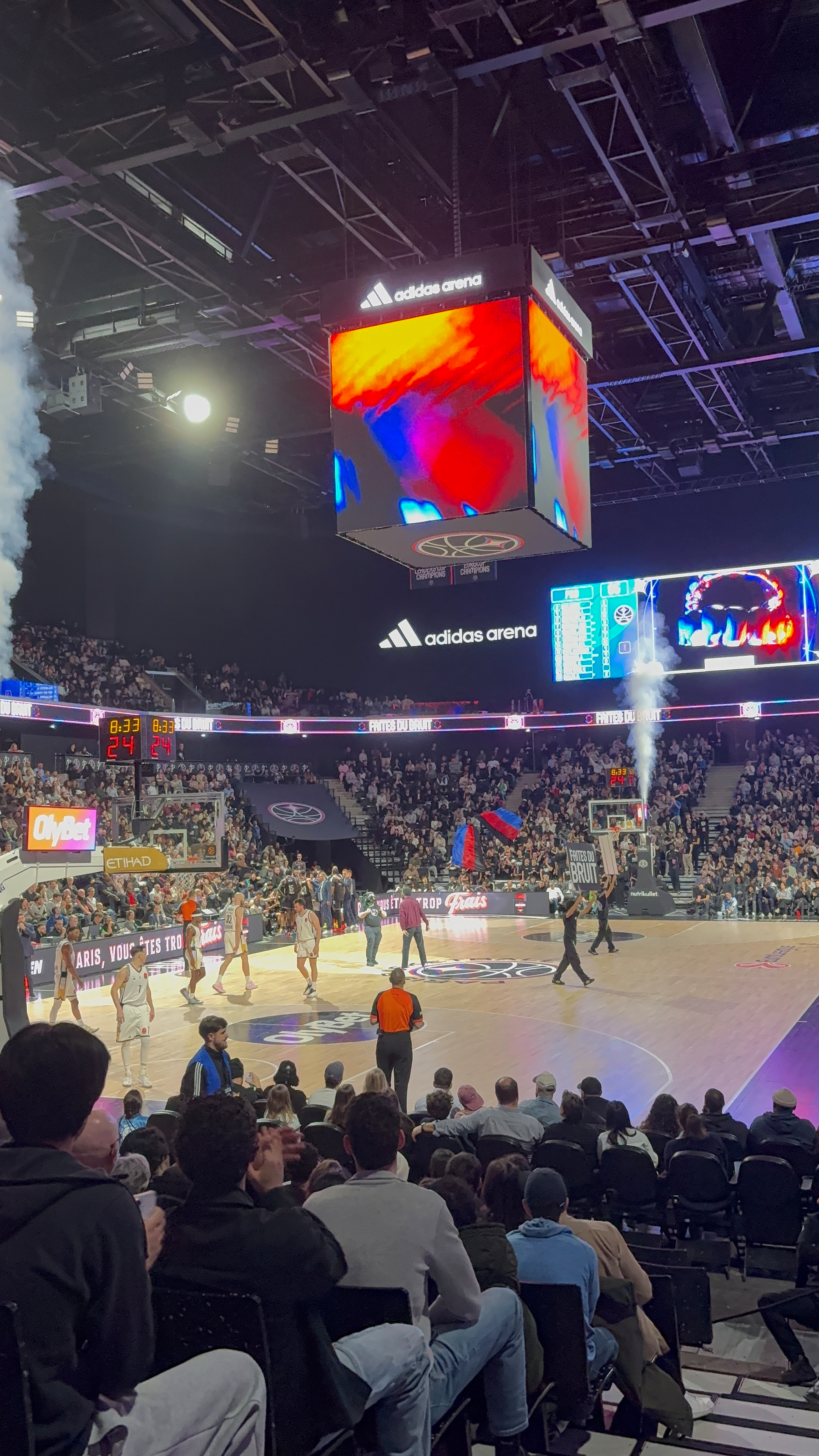 Basketball players are on a court in a packed arena with a large overhead screen displaying visuals.