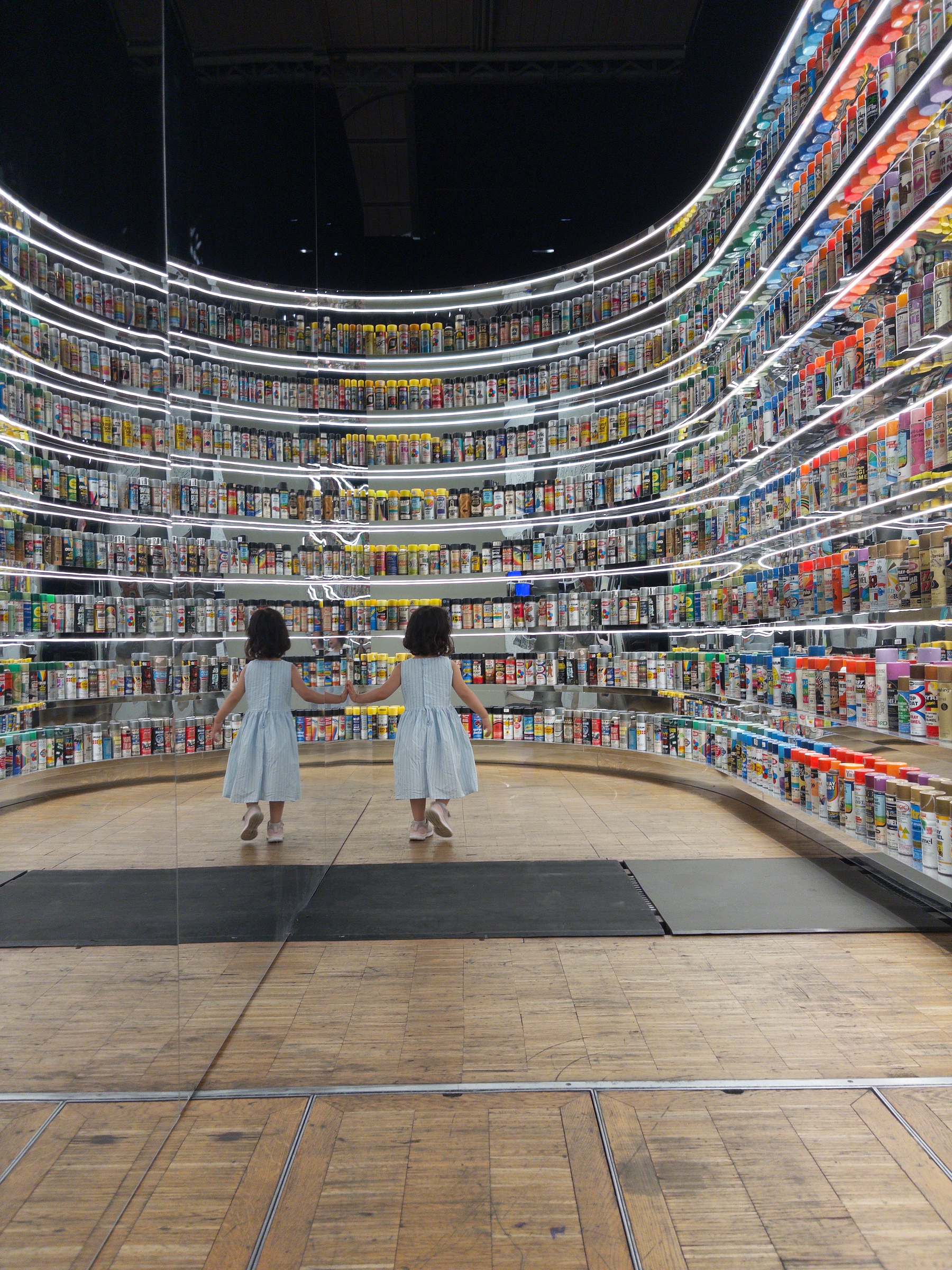 A spacious art gallery features various contemporary artworks, including a colorful mural, whimsical cartoon-like characters on a display, and a black and white patterned installation that visitors are observing.