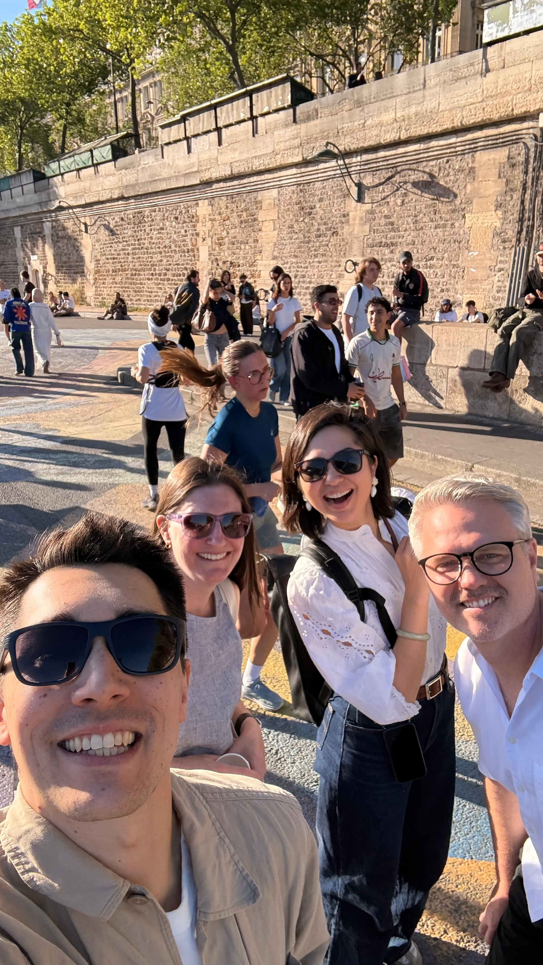 A group of people are smiling and posing for a selfie outdoors on a sunny day with others in the background.